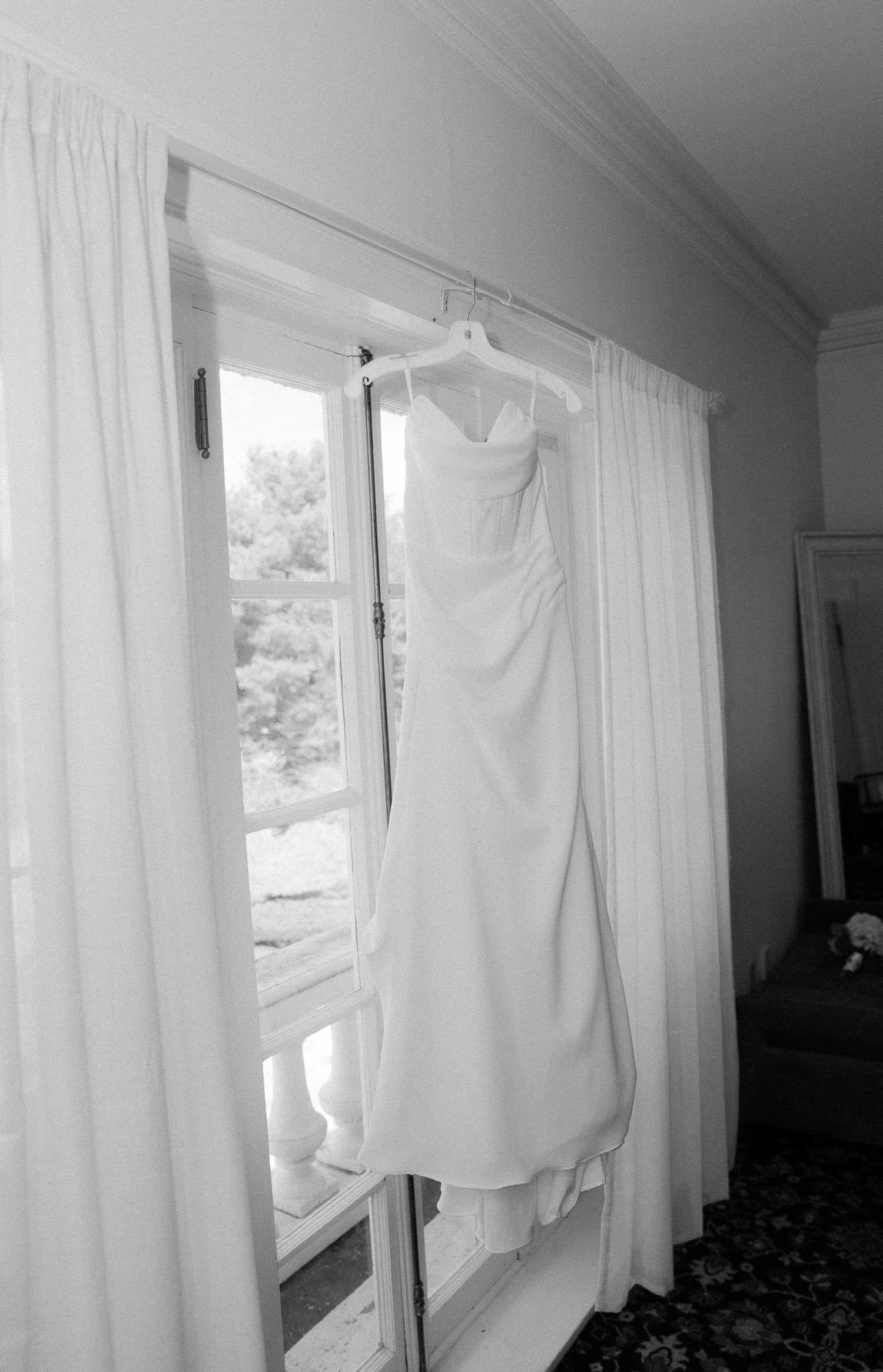 Black and white photo of an elegant white wedding dress hanging on a door frame in front of a bright window.