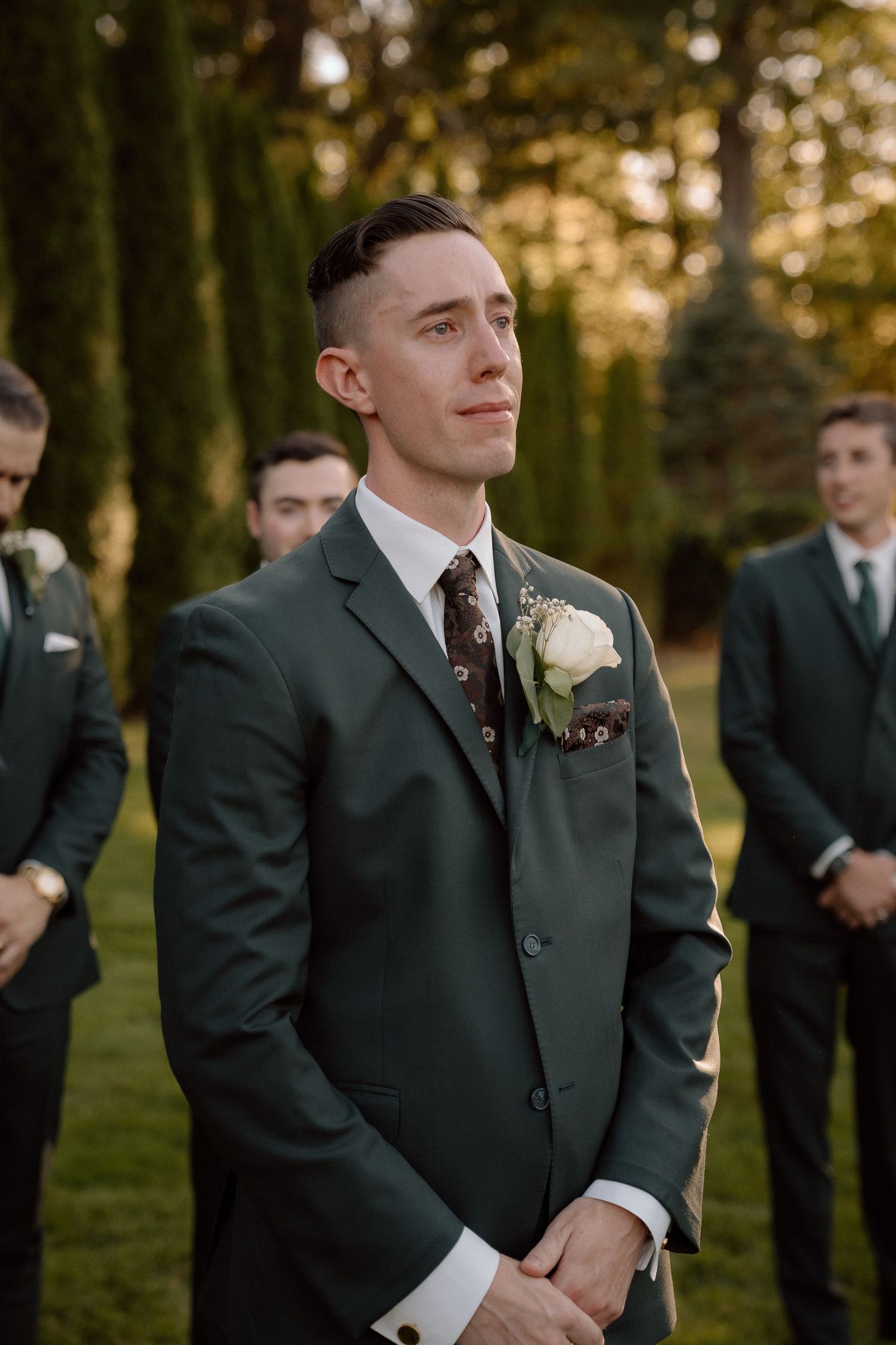 Groomsmen in black suits await the ceremony in warm evening light.