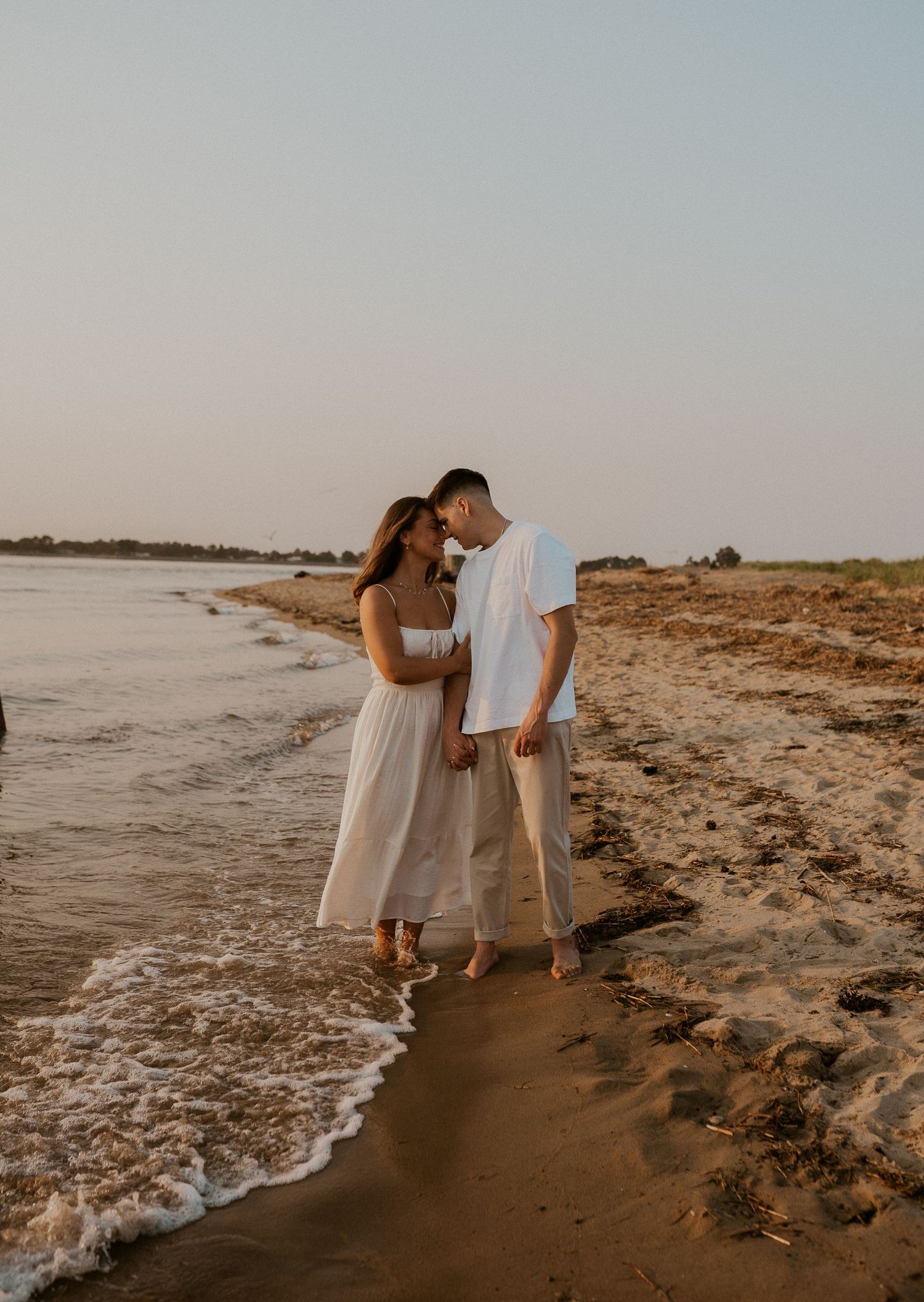 Couple in coordinated outfits poses romantically by the ocean waves at sunset.