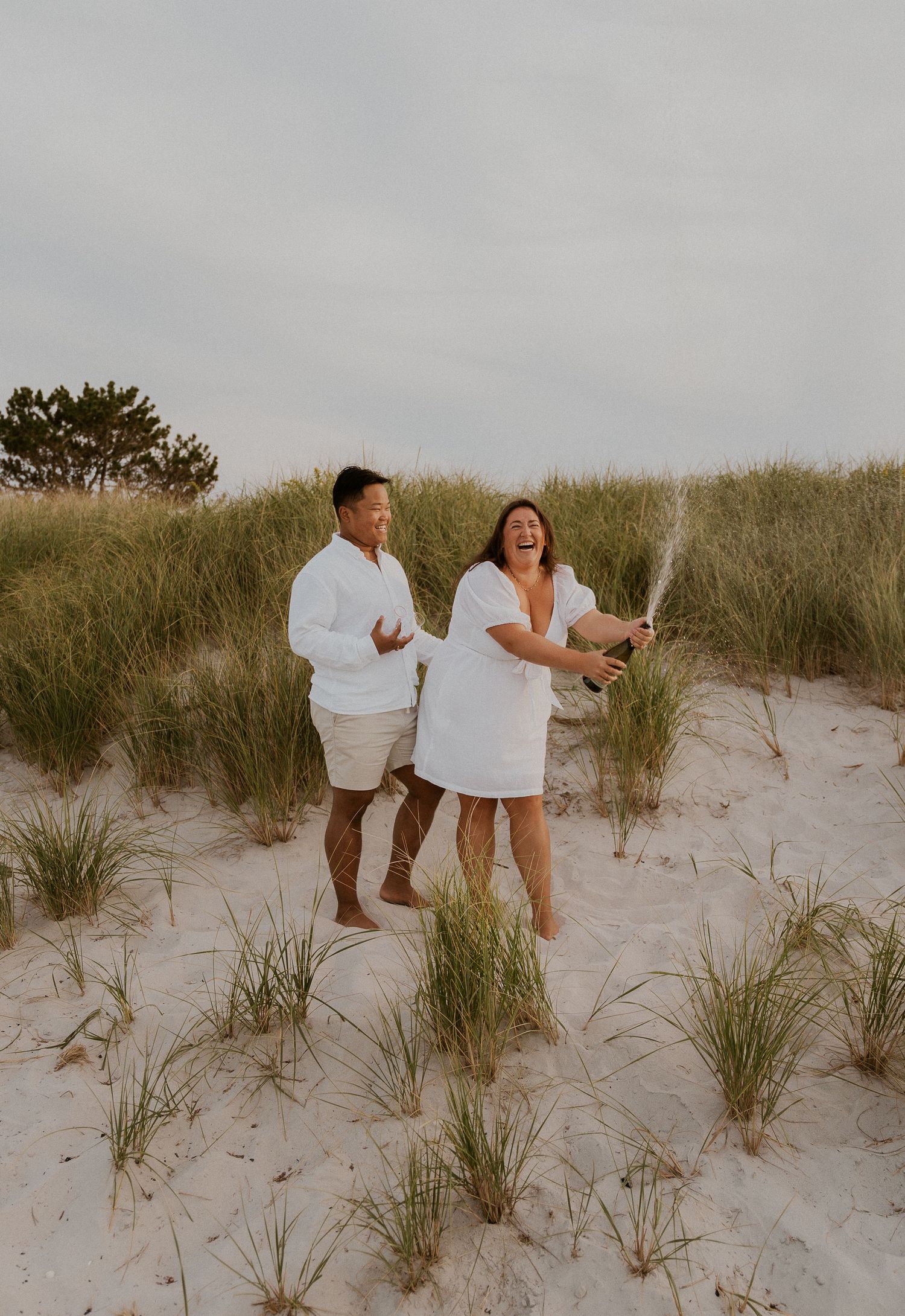Couple sharing playful moments together on sandy beach dunes with tall grass at sunset.
