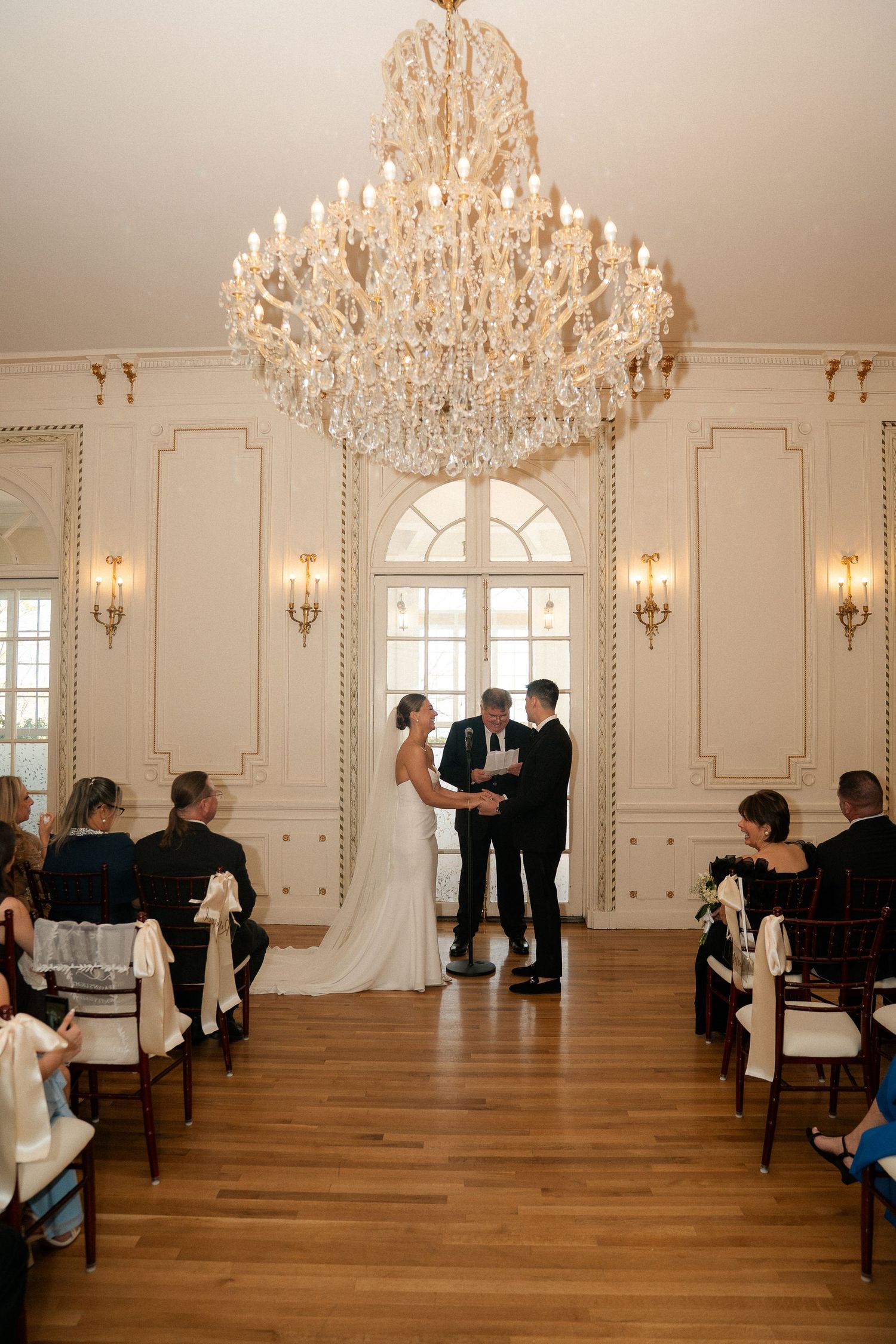 Elegant wedding ceremony in a grand ballroom with crystal chandelier, couple exchanging vows before guests seated in wooden chairs.