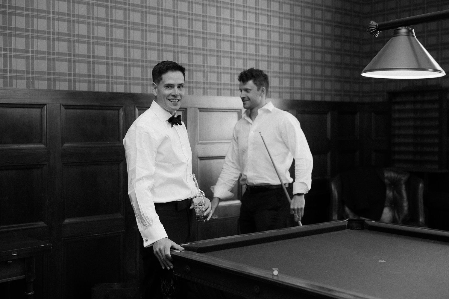 Two men in tuxedos smiling near a billiard table in an elegant wood-paneled room, black and white photo.