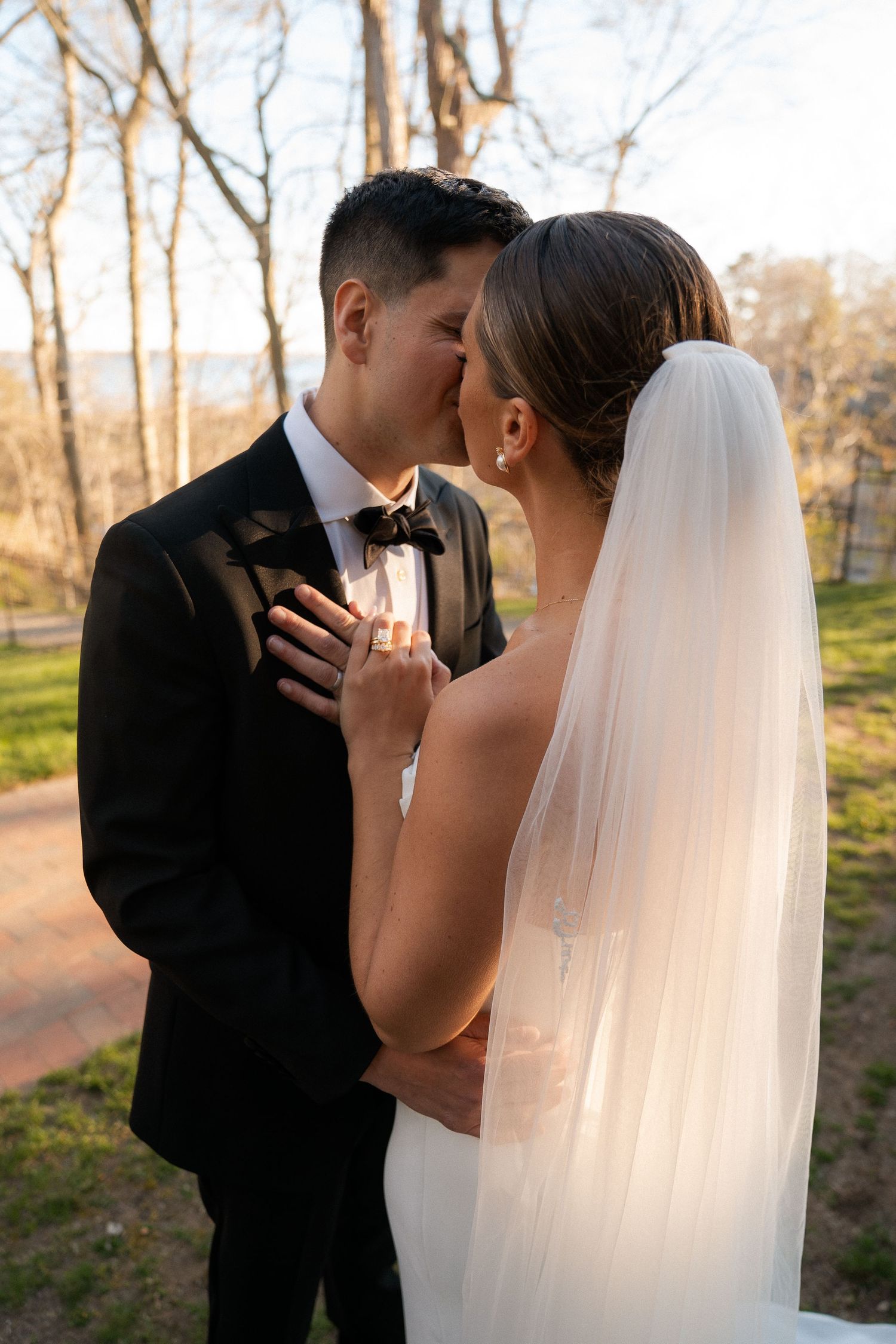 Groom kissing bride on forehead at golden hour, bride in backless wedding gown with cathedral veil.