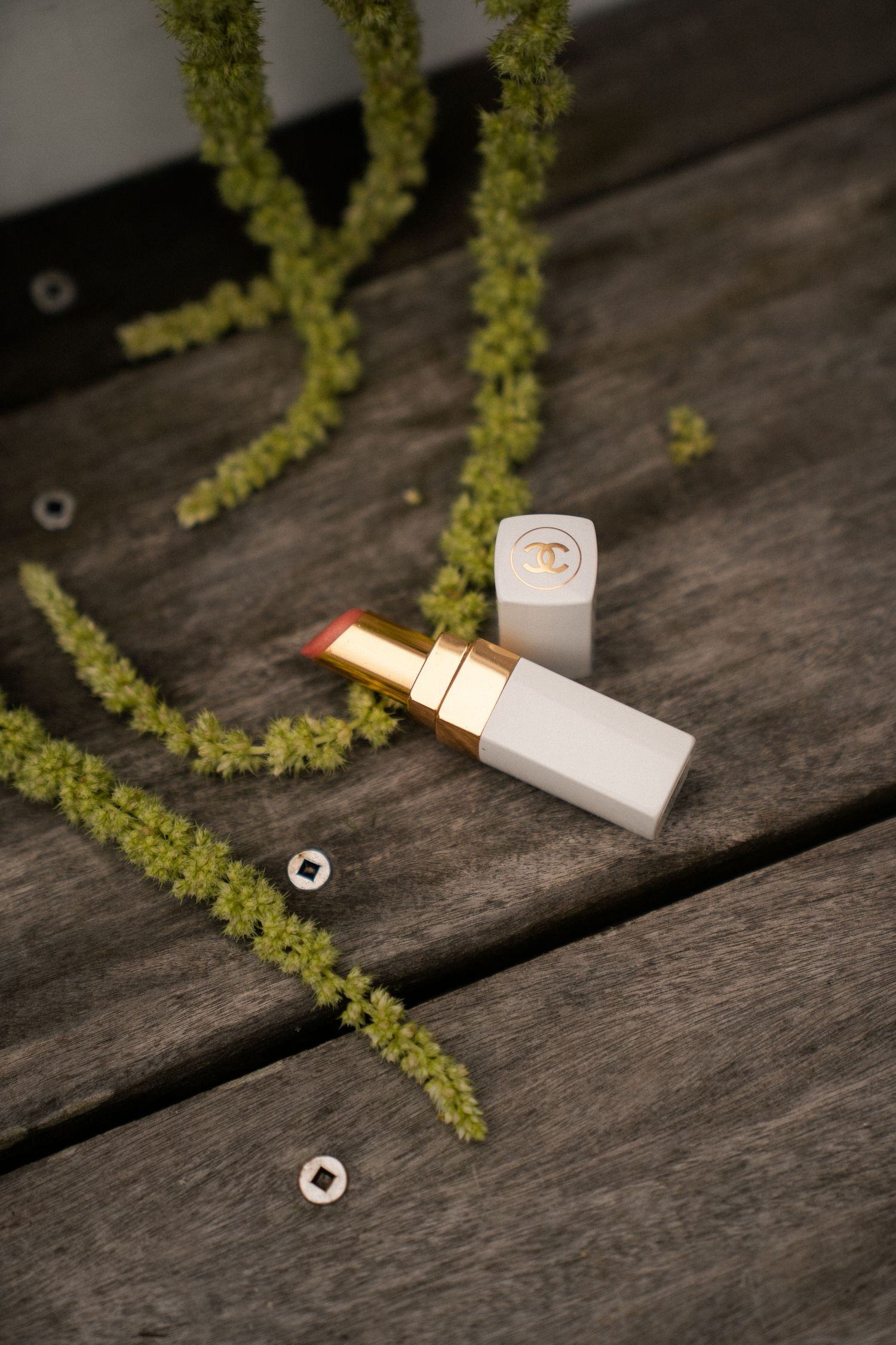 Chanel white lipstick with gold accents open on rustic wood surface with green botanical sprigs.