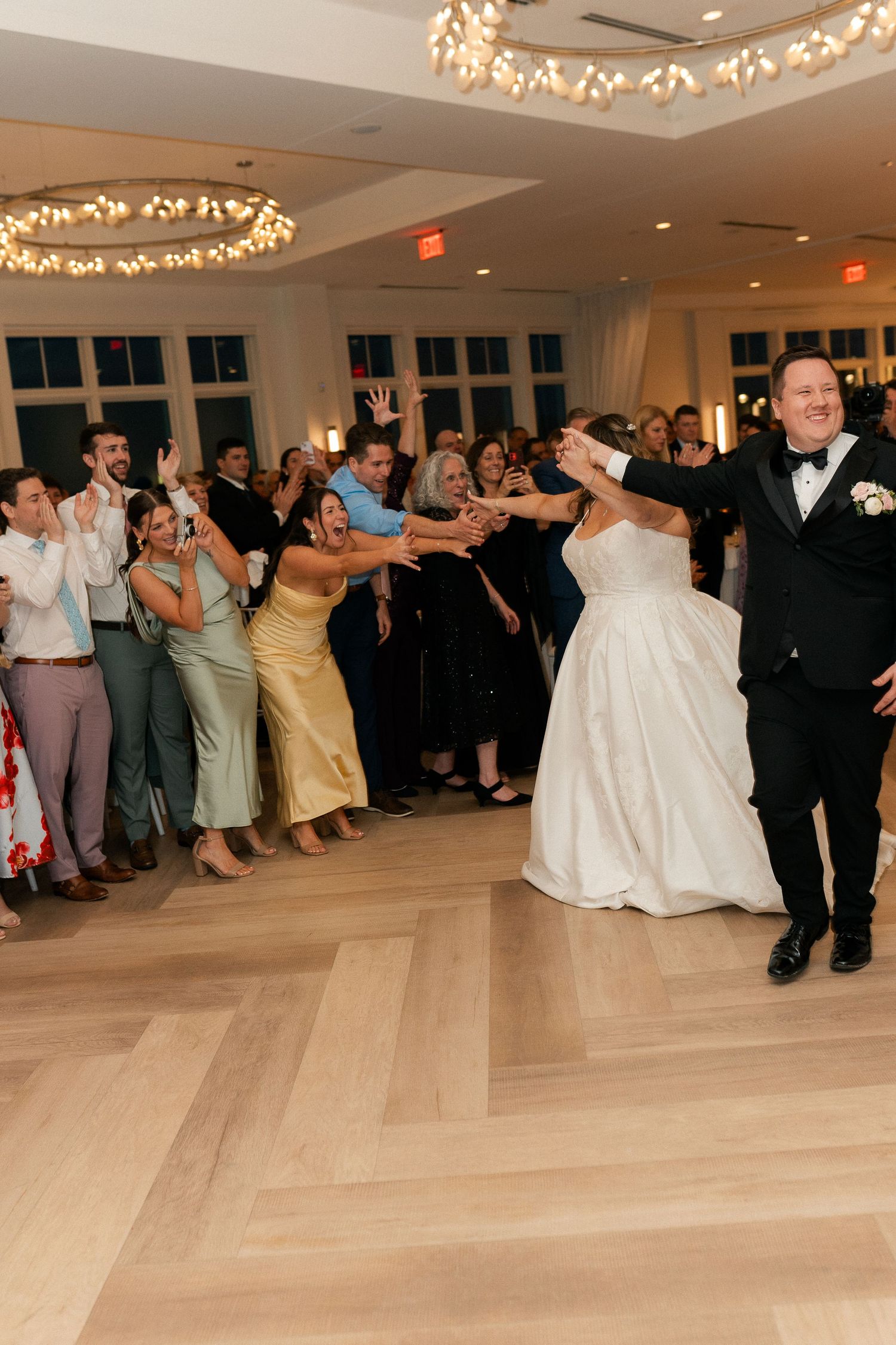 Bride and groom dance with guests during lively wedding reception on herringbone wood floor.