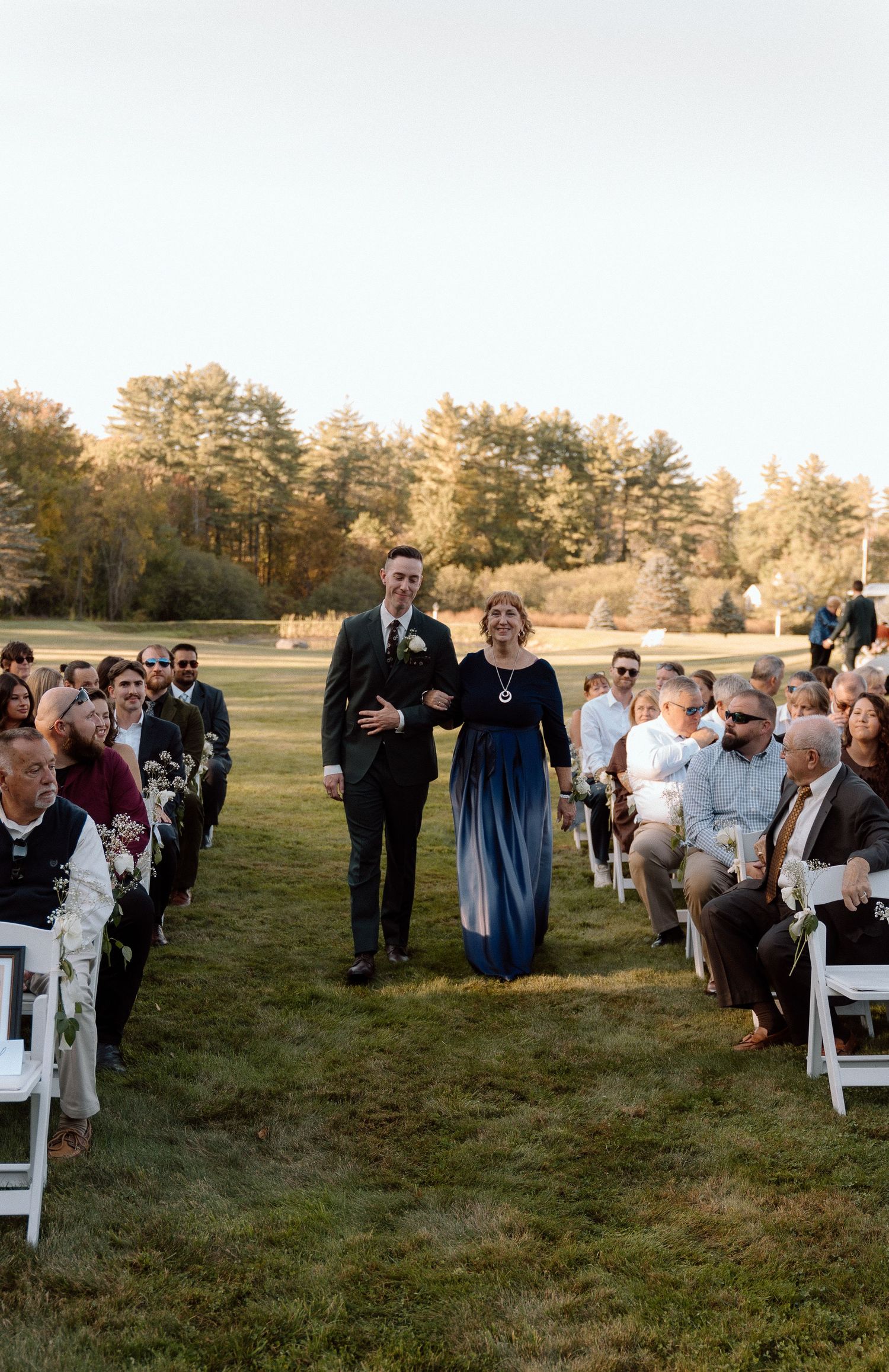 Wedding guests seated in white chairs during an outdoor autumn ceremony.