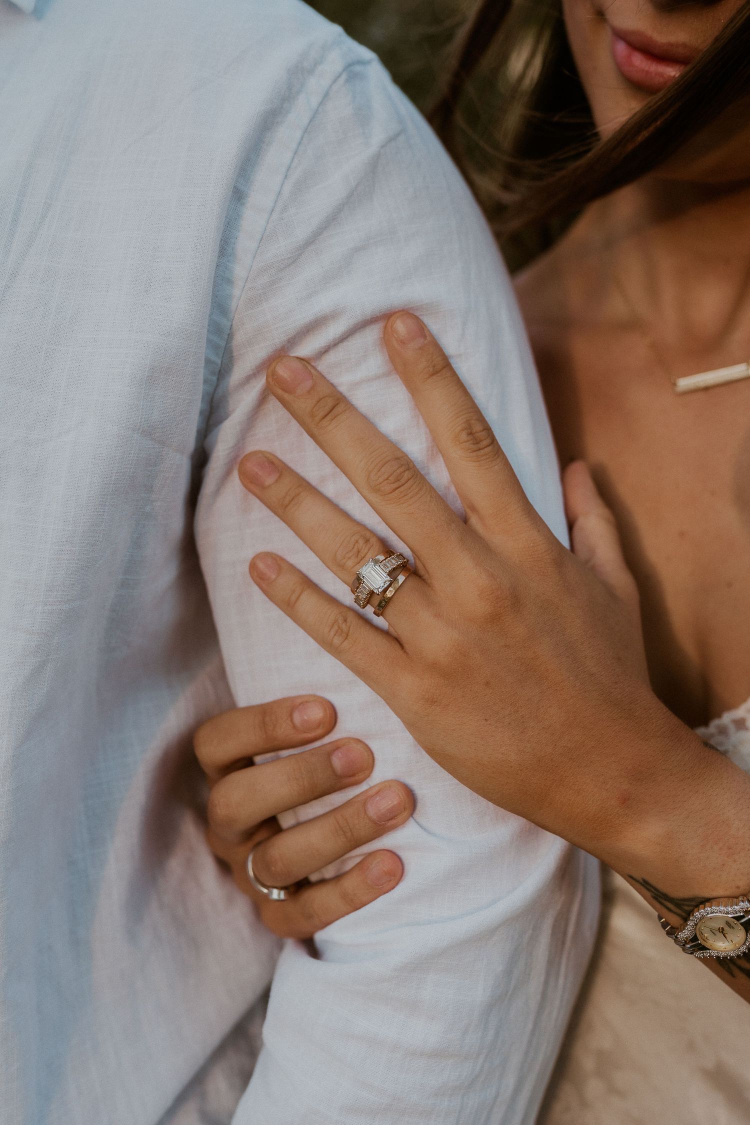 Close-up of a woman's hand with an elegant emerald-cut engagement ring, resting on a man's arm.