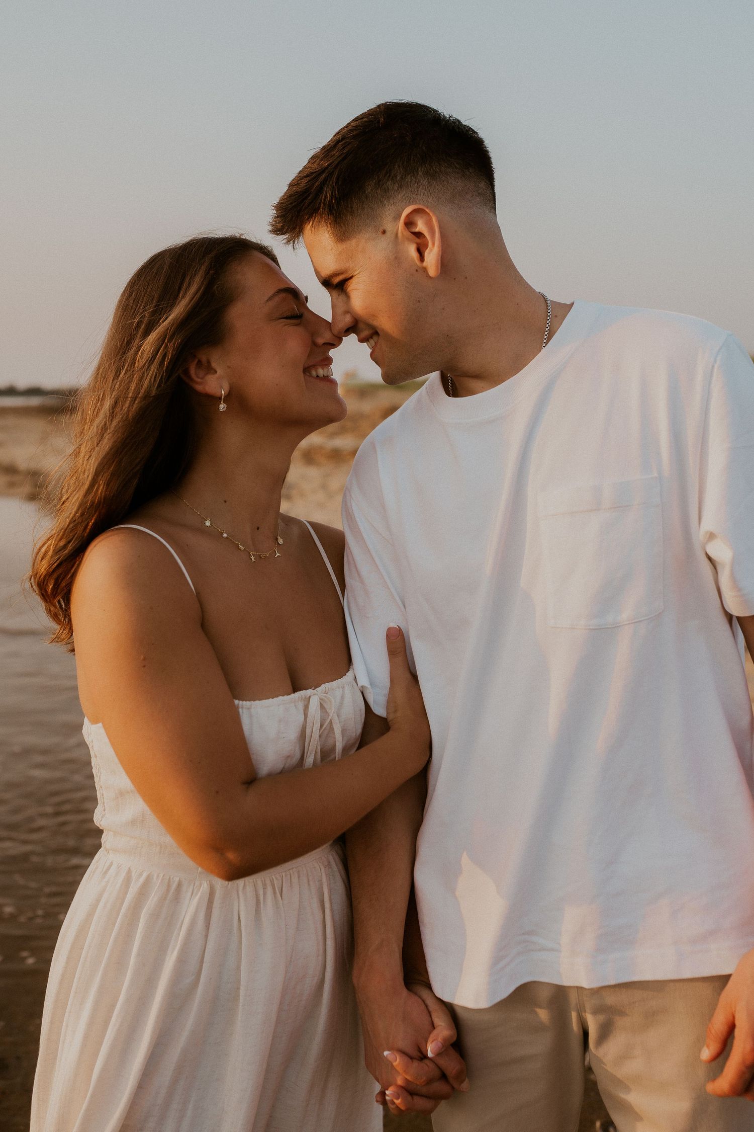 Intimate couple shares tender moments on a beach during sunset wearing matching white outfits.