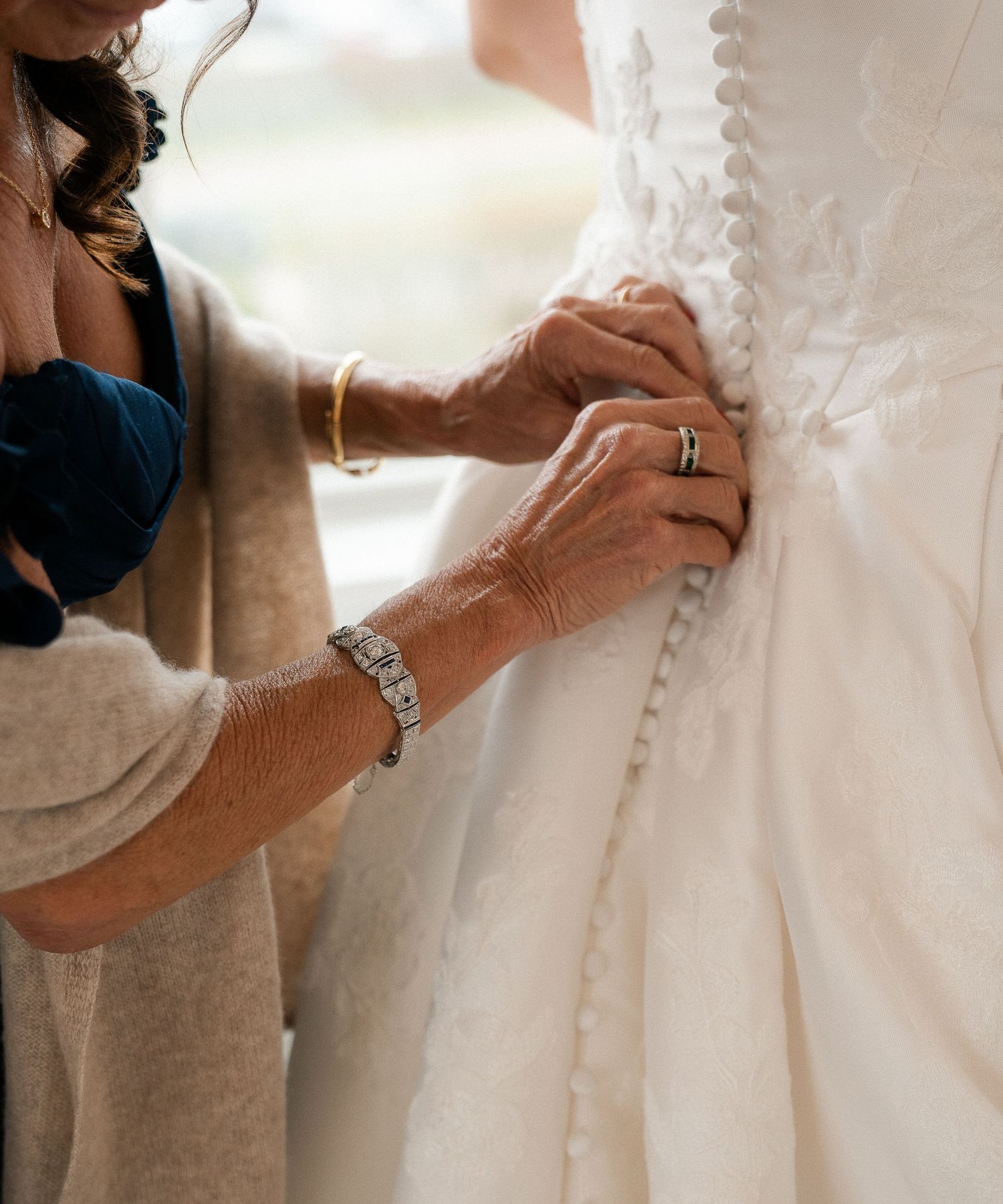 Mother carefully fastening delicate button closures on the back of a bride's lace wedding gown.