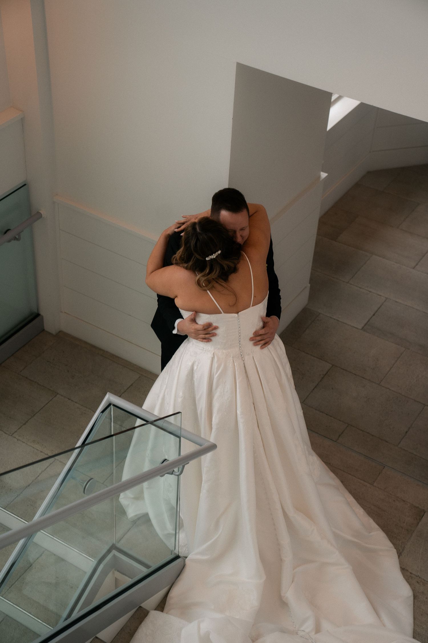 Bride and groom sharing emotional first look hug on staircase, bride wearing elegant white ball gown.