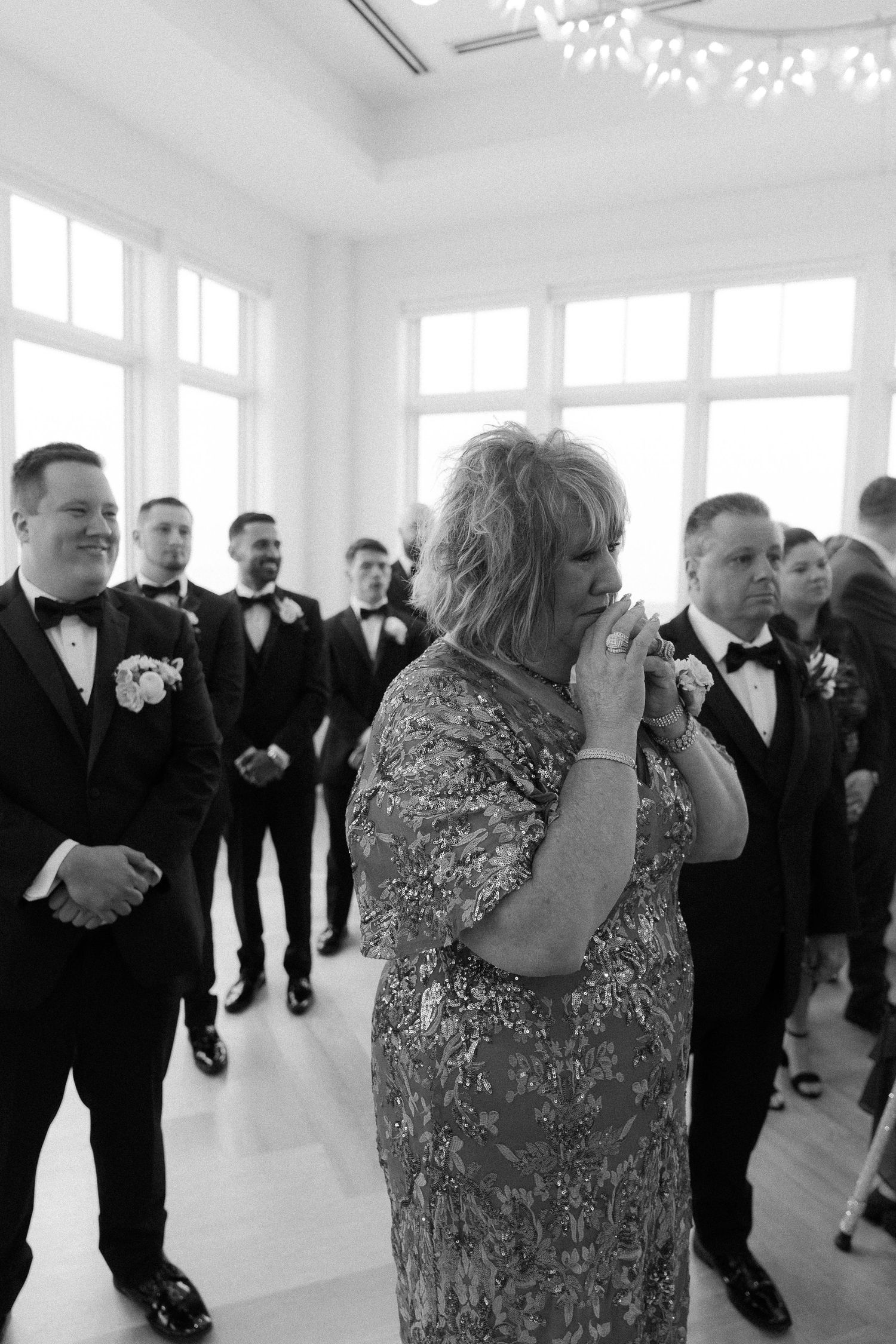 Emotional mother of the groom clasping hands during wedding ceremony surrounded by groomsmen in black tuxedos.