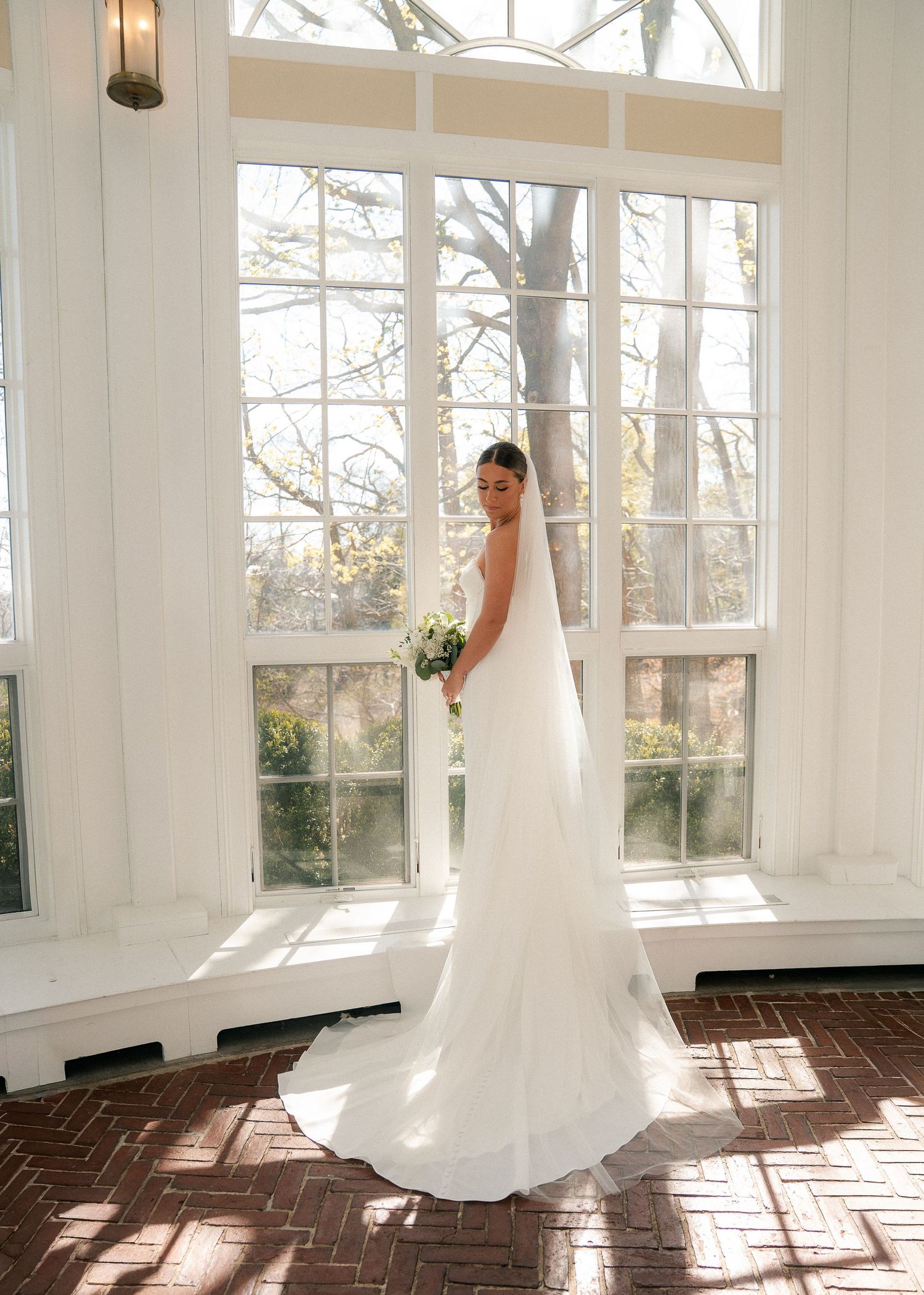 Bride in white gown and veil holding bouquet stands by large sunlit windows on brick floor.