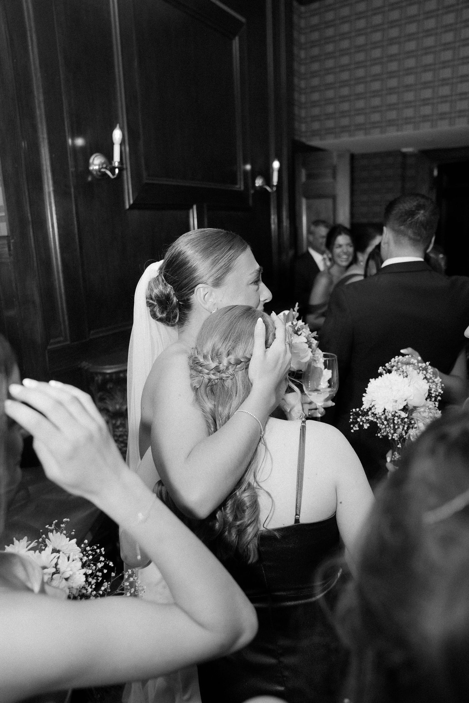Black and white photo of a bride in veil embracing a guest at a wedding reception, holding bouquets.