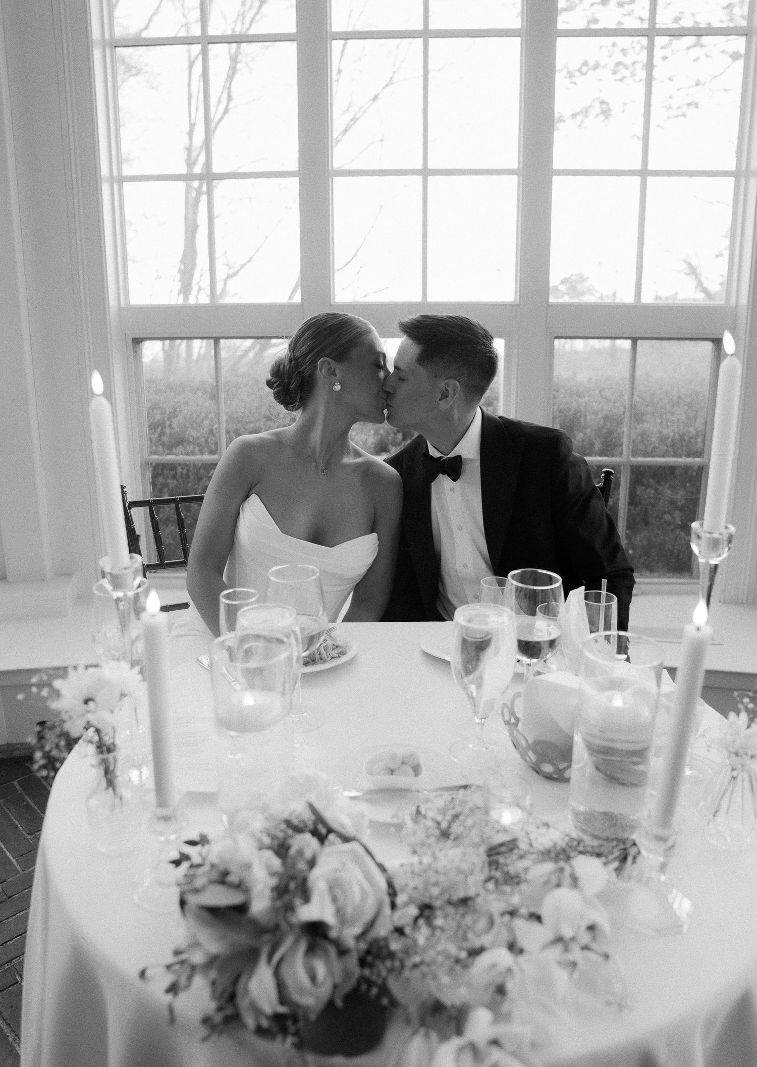 Black and white photo of newlyweds kissing at sweetheart table with candles and floral centerpiece.