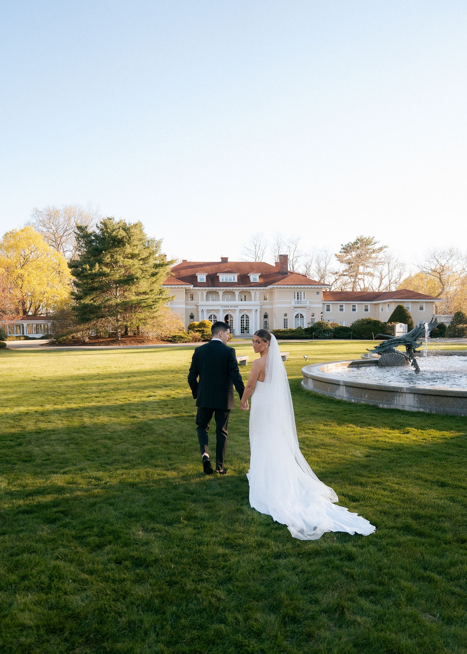 Bride and groom walk hand in hand across lush green lawn toward elegant mansion with fountain at golden hour.