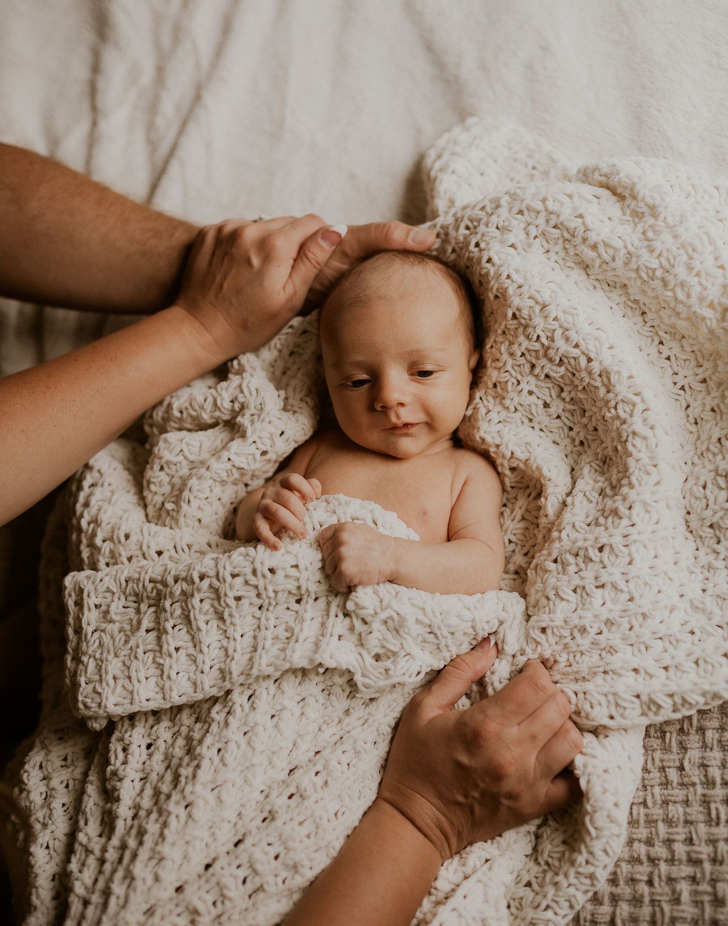 Baby wrapped in a knit blanket smiling while being touched by caring hands.