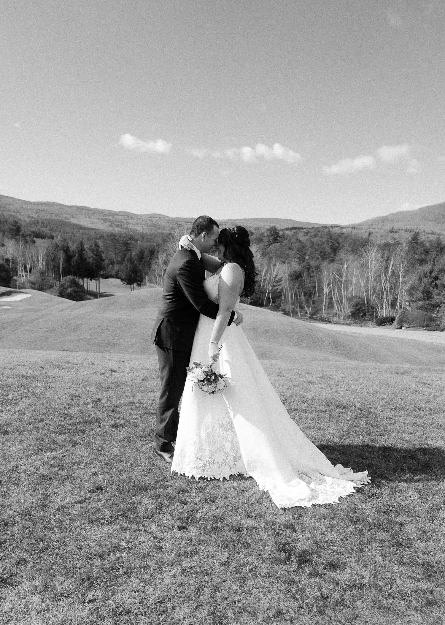 A romantic black and white wedding photo of newlyweds kissing on a hilltop with mountain views.