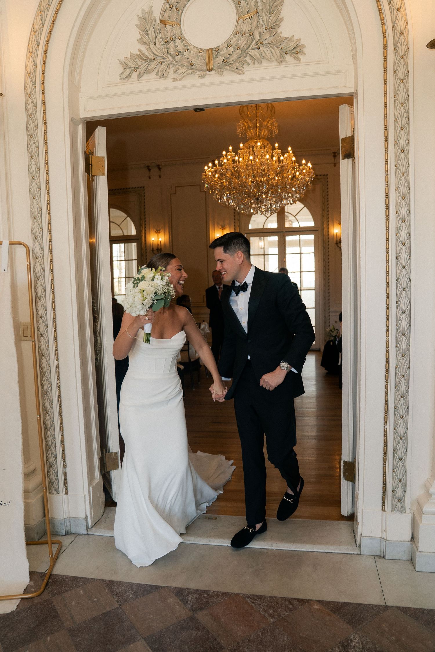 Bride and groom holding hands walking through elegant doorway with chandelier at wedding reception.