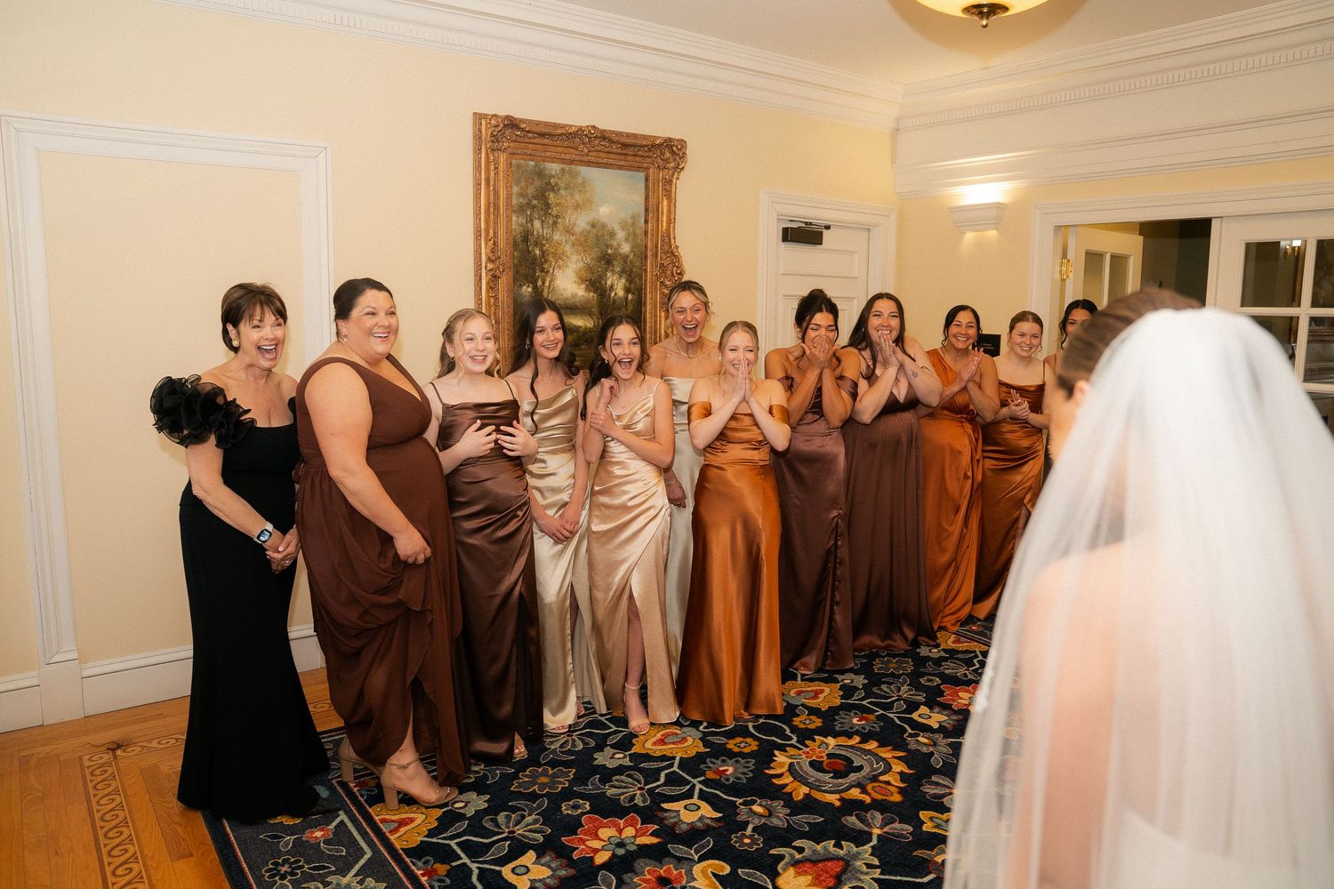 Bridesmaids in brown and copper satin gowns react with joy as bride in white veil reveals her wedding look.