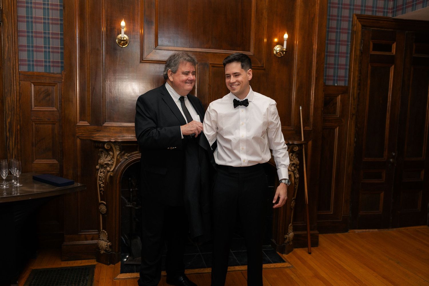 Two men in formal tuxedos smiling together in an elegant wood-paneled room with a fireplace.