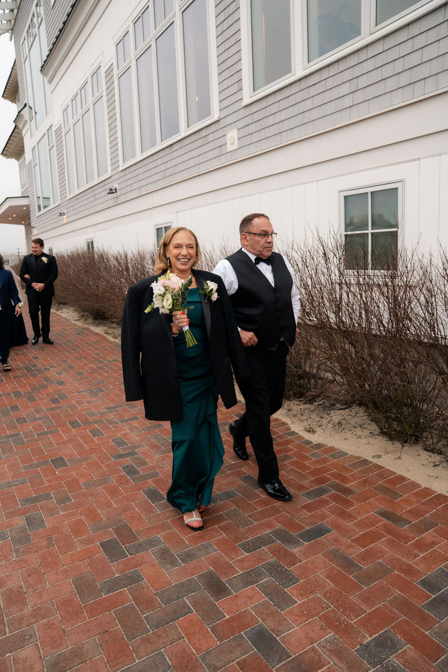 Bridesmaid in emerald green gown and black jacket walks with groomsman in tuxedo on brick path outside wedding venue.