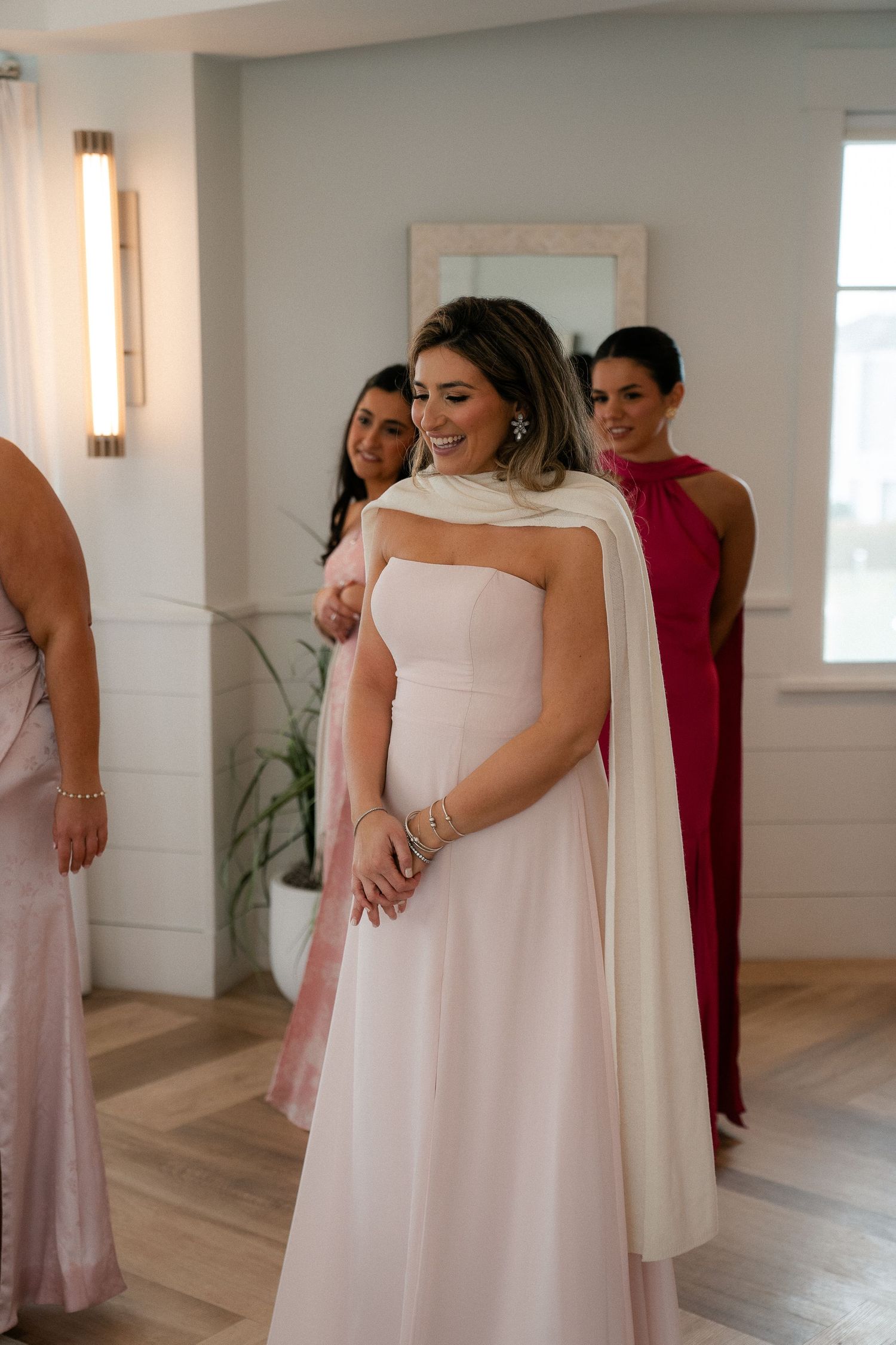 Bride in blush strapless gown with white cape smiles as bridesmaids in pink and red dresses look on.