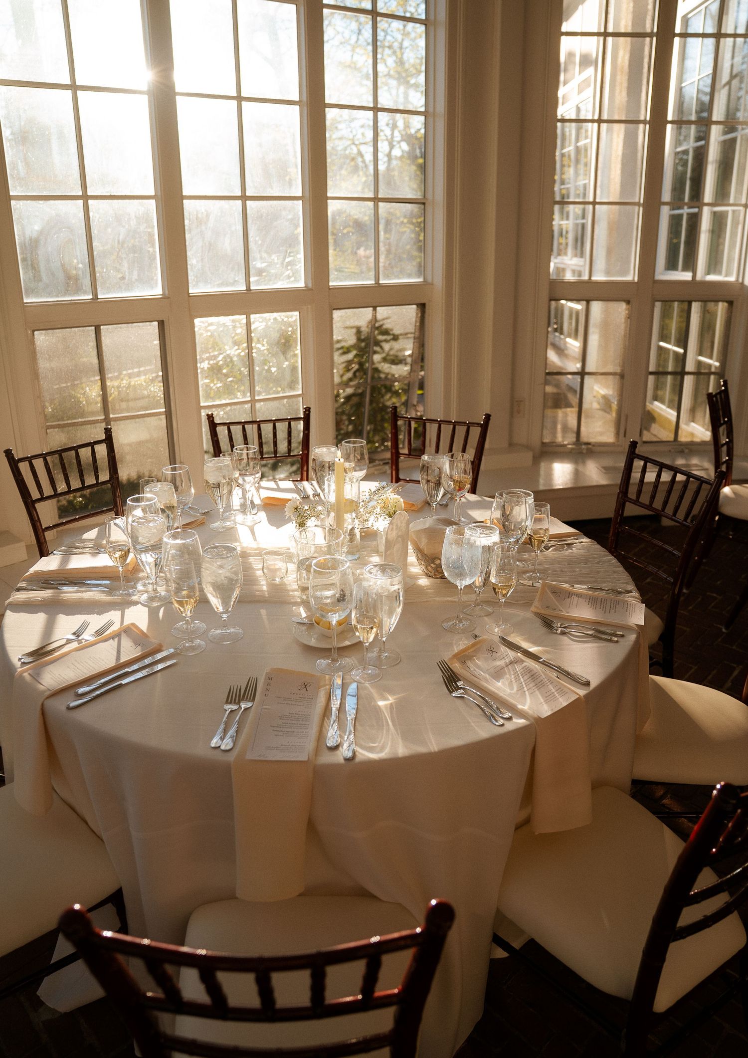 Elegant round banquet table set with white linens, crystal glassware, and candles in a sunlit ballroom with tall windows.
