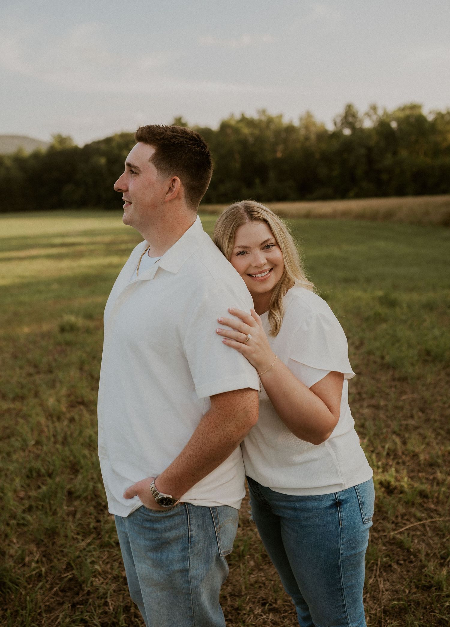 Couple in matching white t-shirts and jeans embrace while standing in a sunlit grassy field at sunset.
