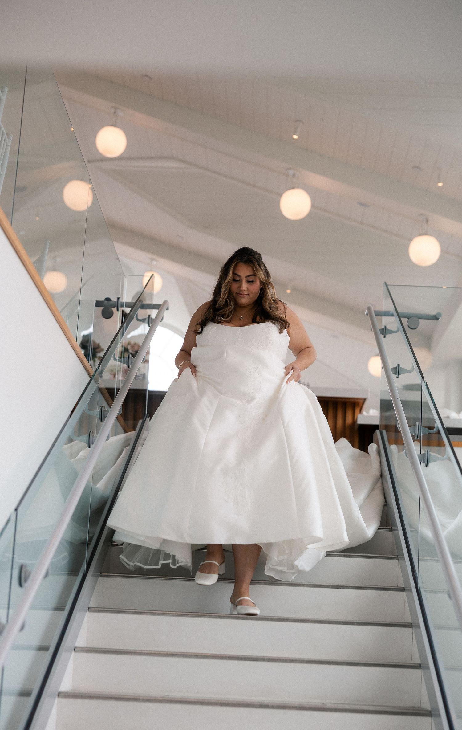 Bride in strapless tea-length ball gown descending modern staircase with globe lights.