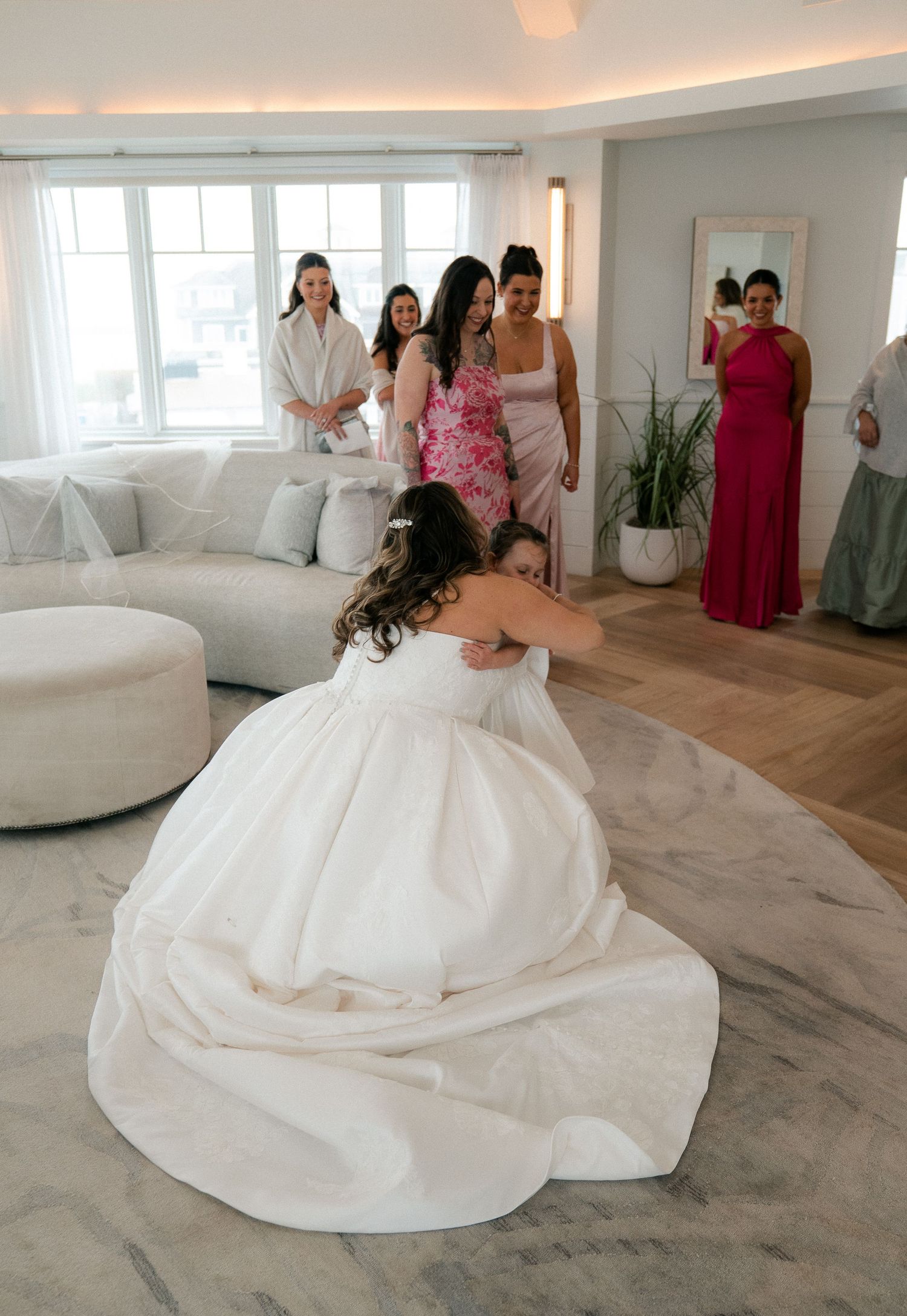 Bride in a white ball gown hugging a young flower girl as bridesmaids look on with smiles.