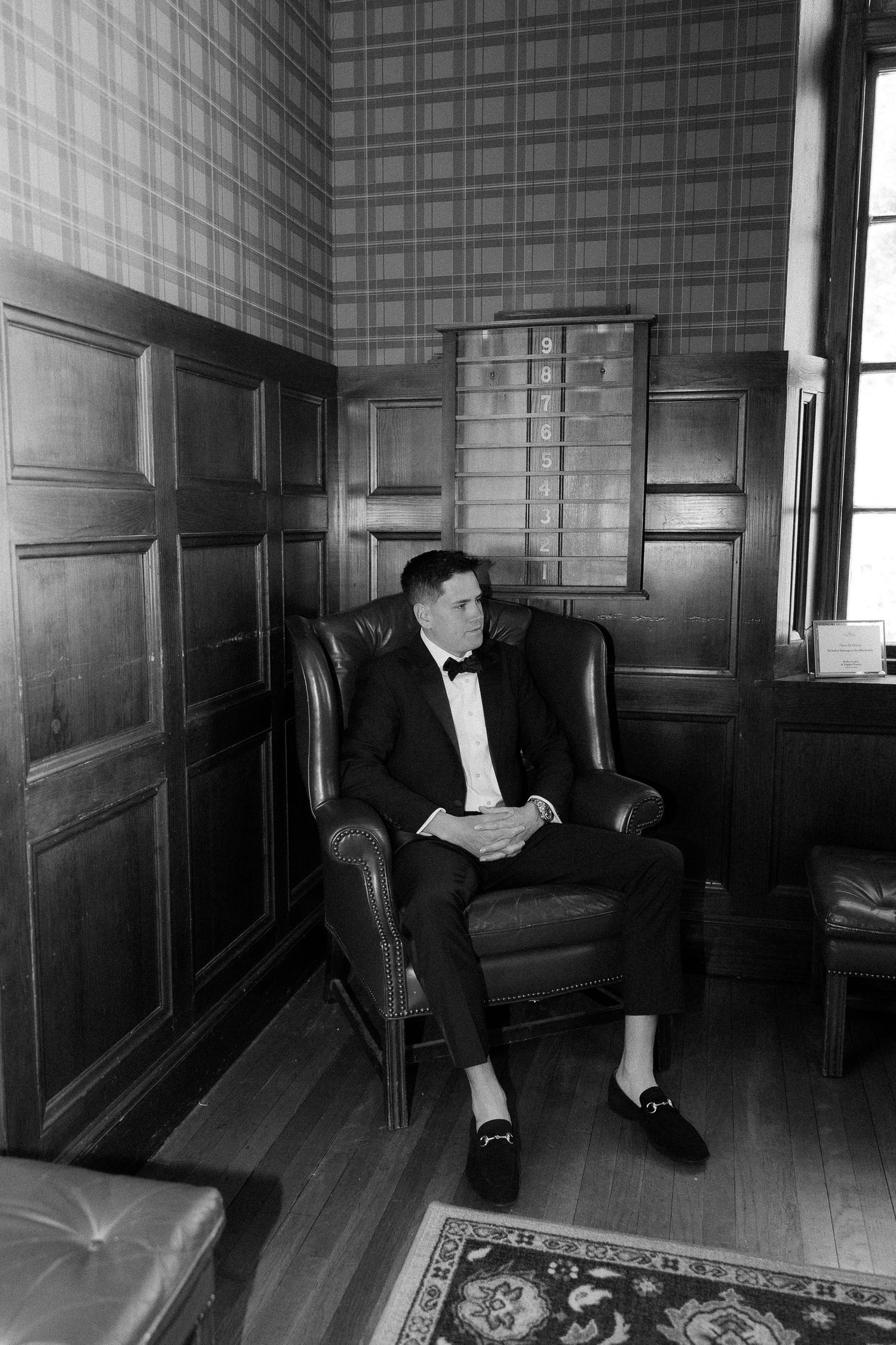 A groom in a tuxedo and bow tie sits in a leather armchair in an elegant wood-paneled room, black and white photo.