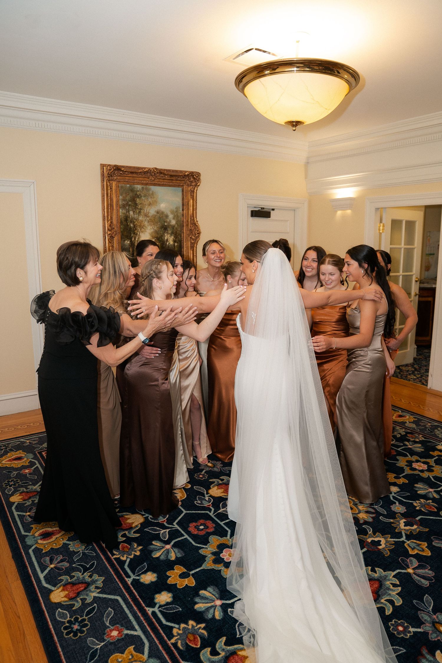 Bride in white gown and veil surrounded by bridesmaids in brown satin dresses during wedding celebration indoors.