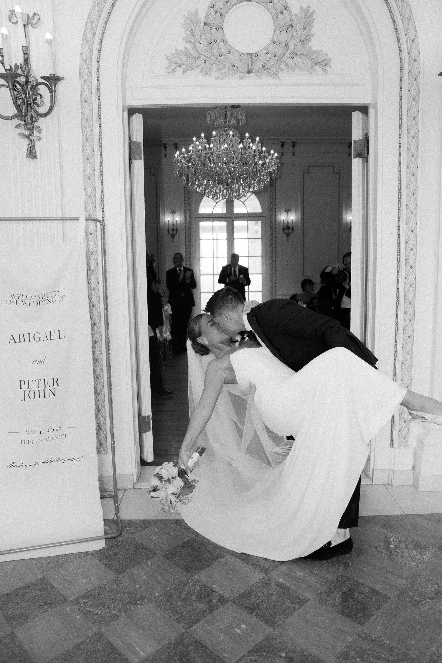 Groom dips bride in a romantic kiss at the entrance of their elegant wedding venue in black and white photo.