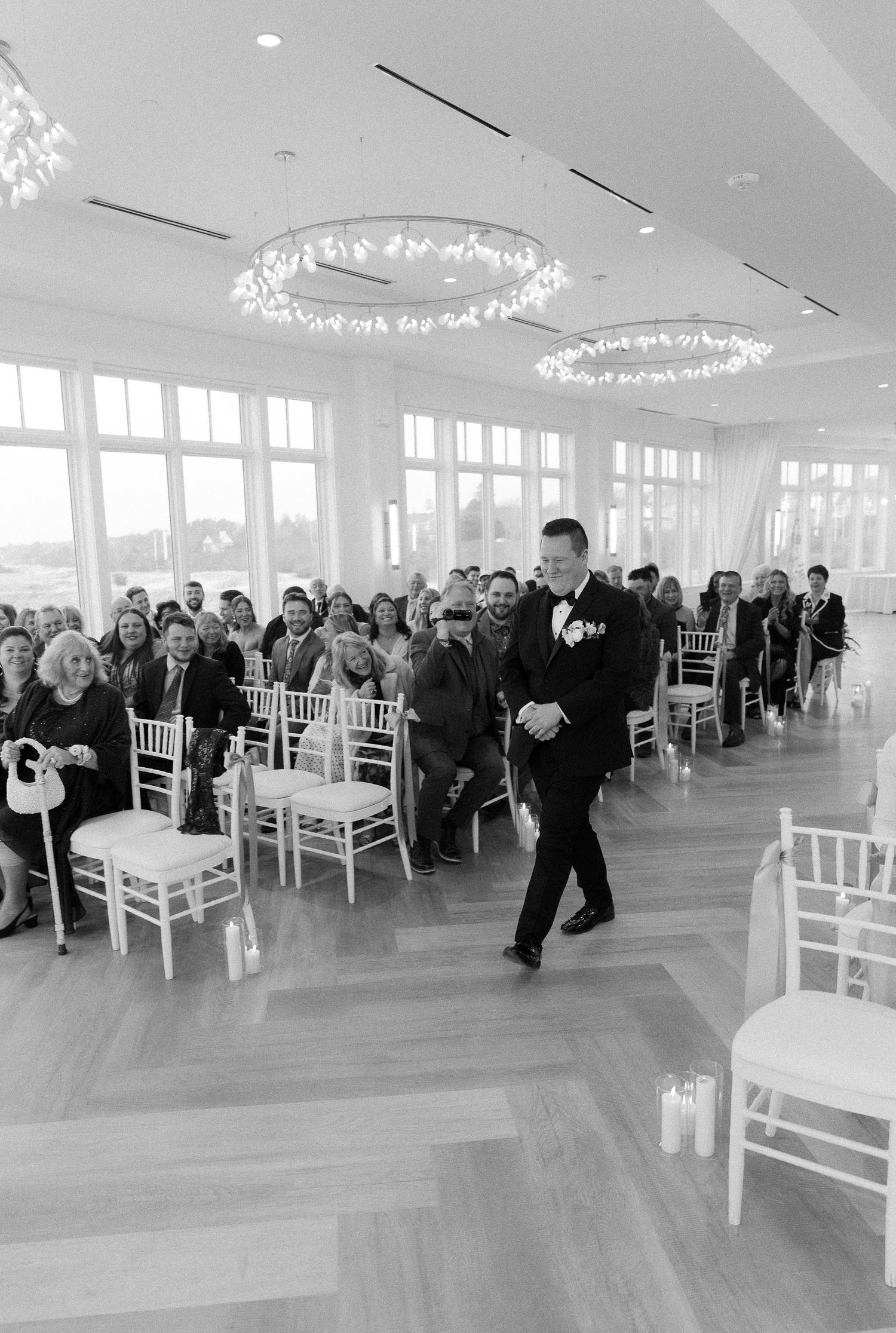 Groom in black tuxedo walks down the aisle of a bright oceanview wedding venue filled with seated guests.