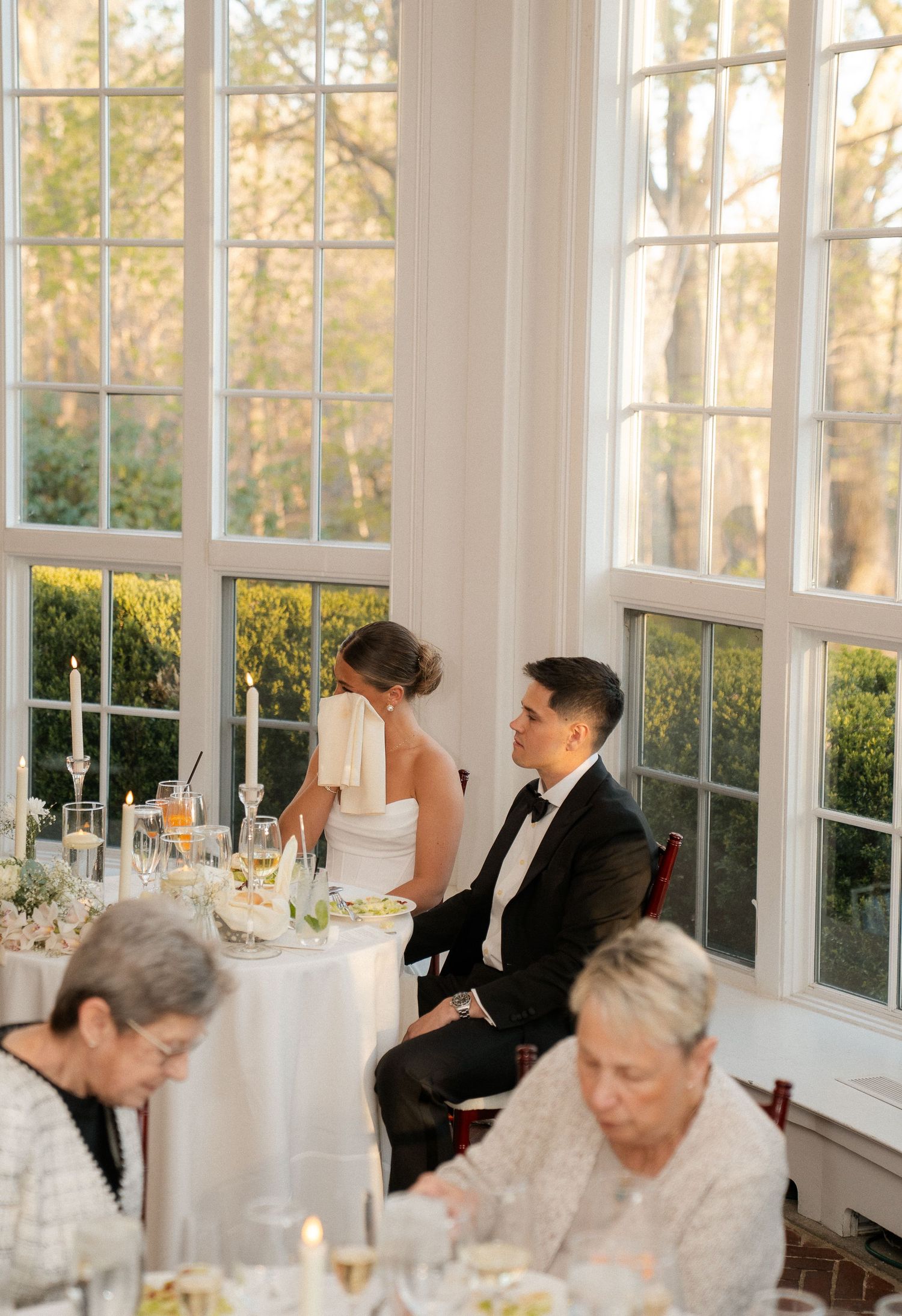 Bride and groom seated at elegant wedding reception table near large windows with garden views.