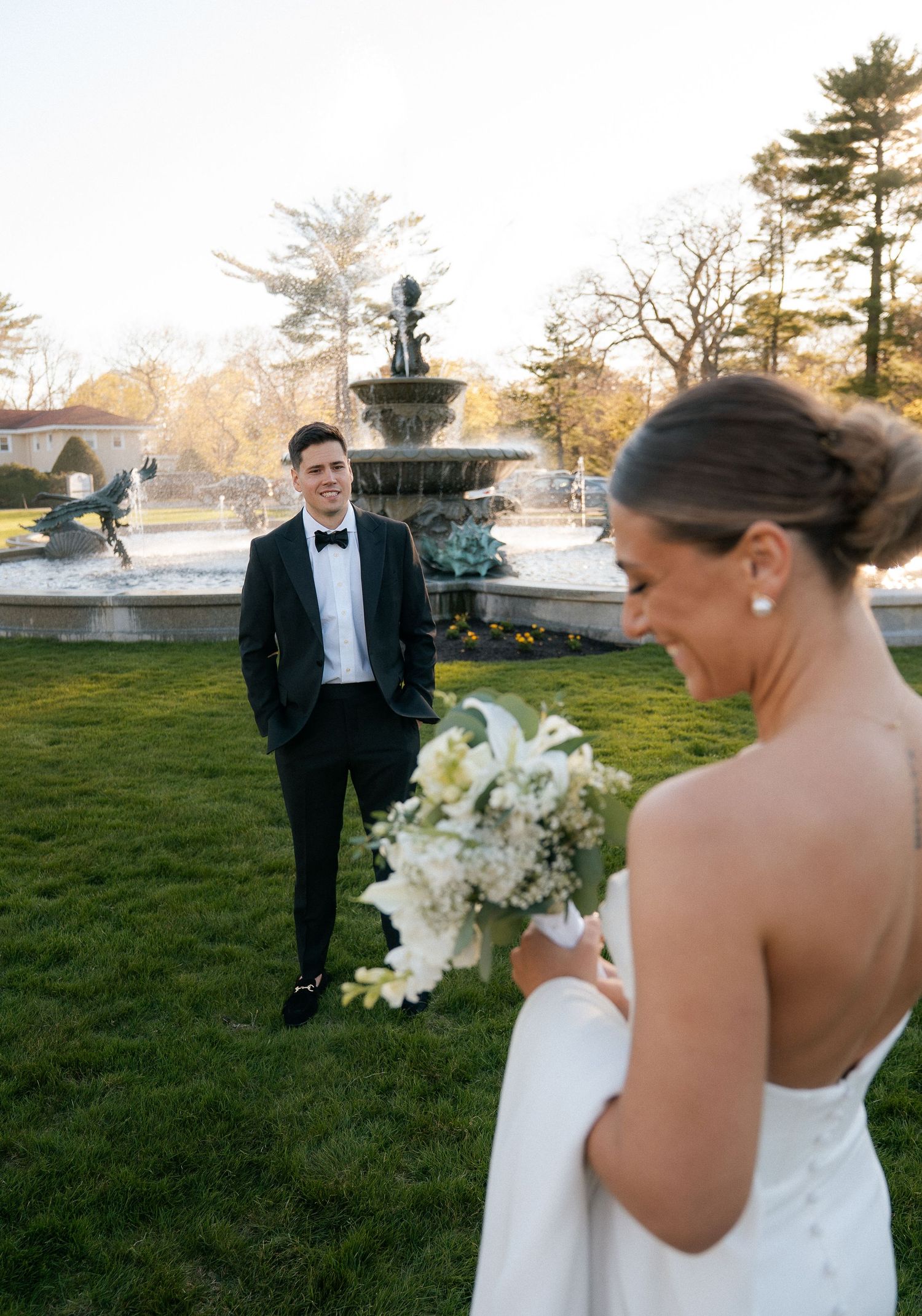 Bride holding white bouquet smiles as groom in black tuxedo stands by ornate fountain at outdoor wedding.