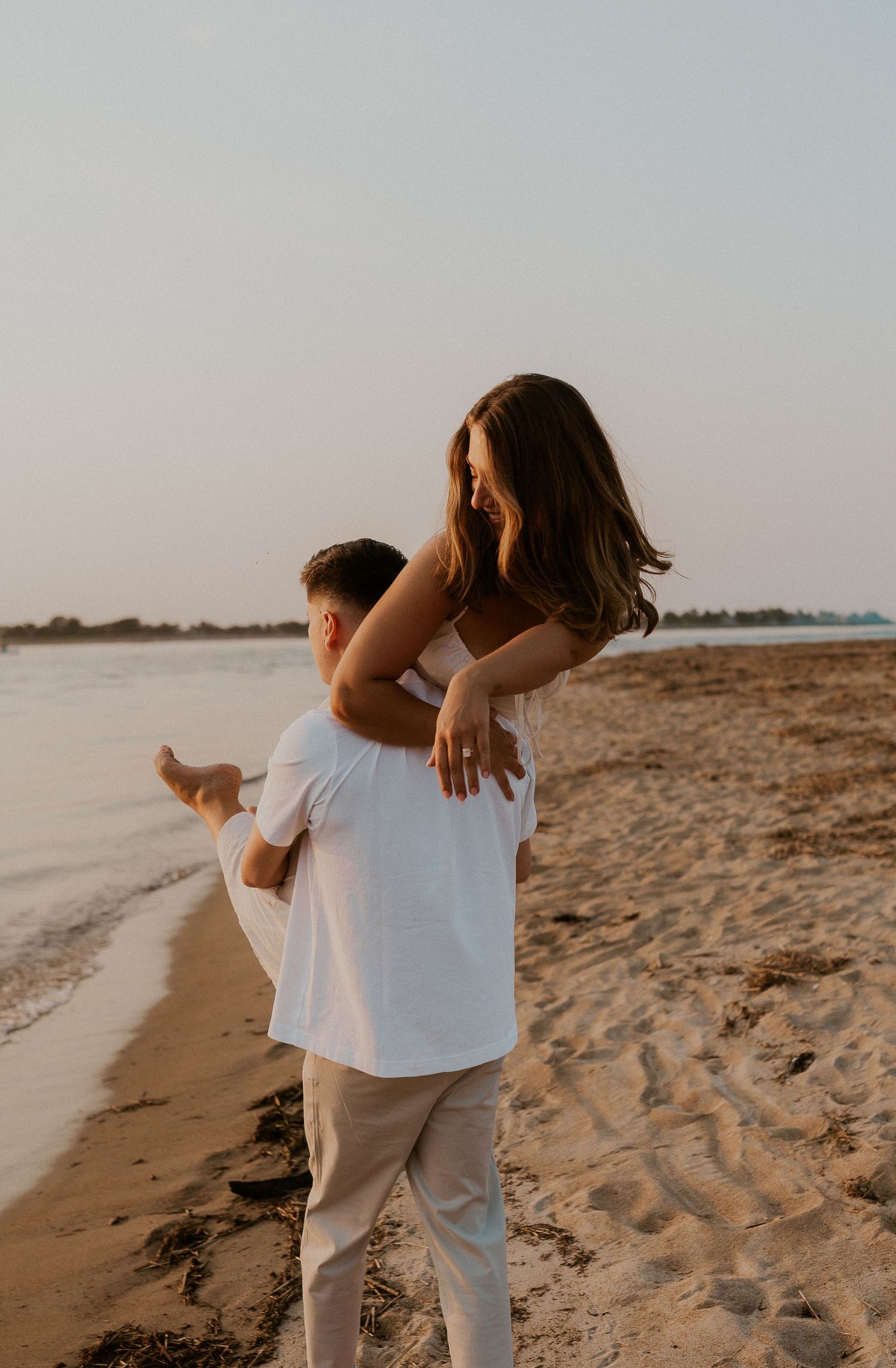 Couple enjoying playful piggyback moment on beach with wooden posts in background.