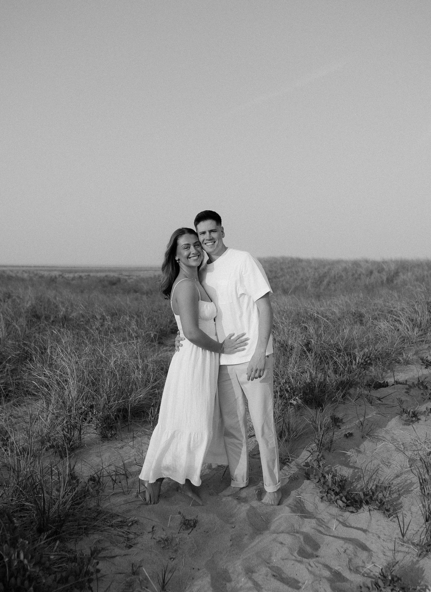 Black and white photo of a couple sharing an intimate moment on sand dunes surrounded by coastal vegetation.