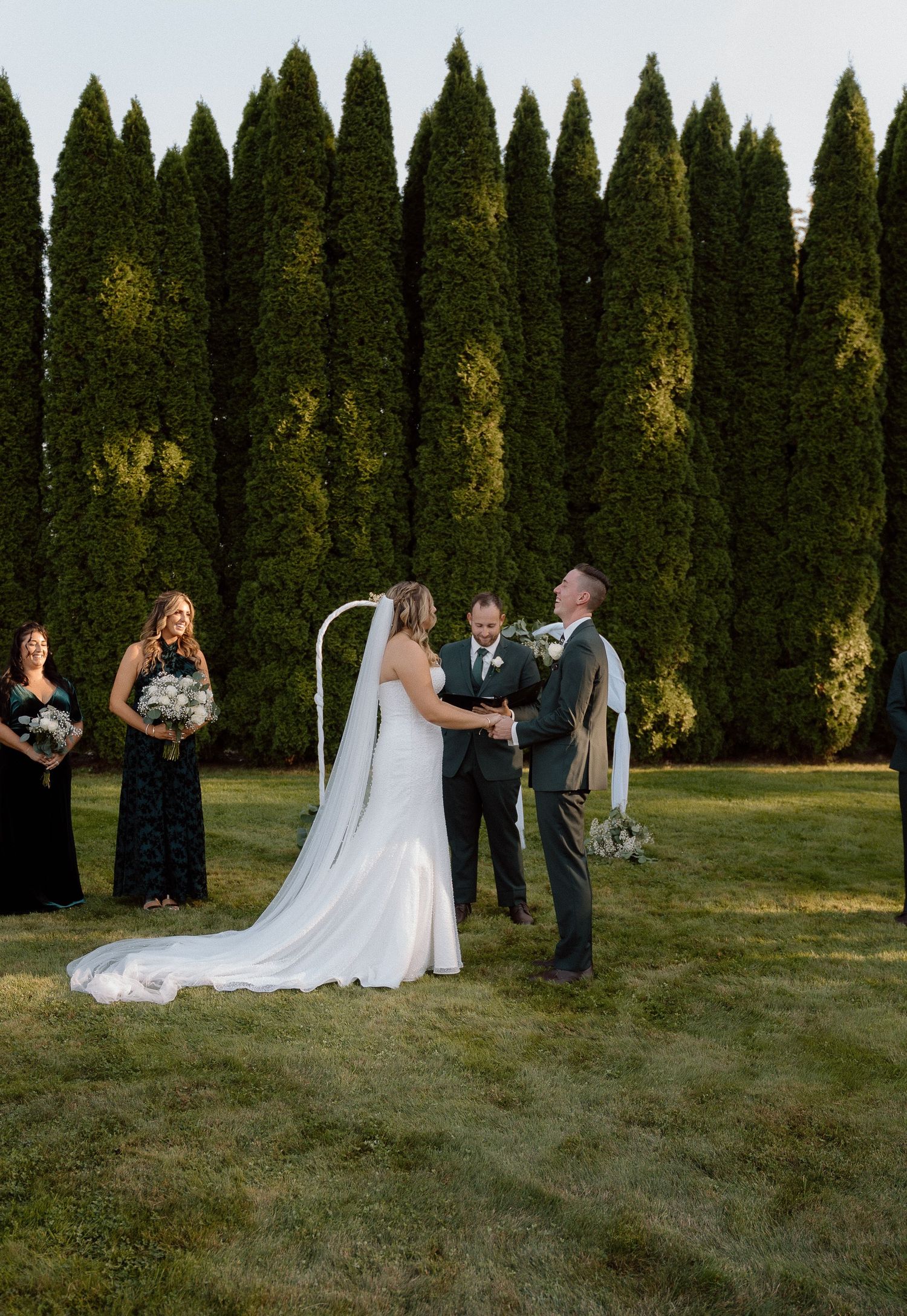 Wedding party gathered on lawn with elegant cypress tree backdrop during outdoor ceremony.