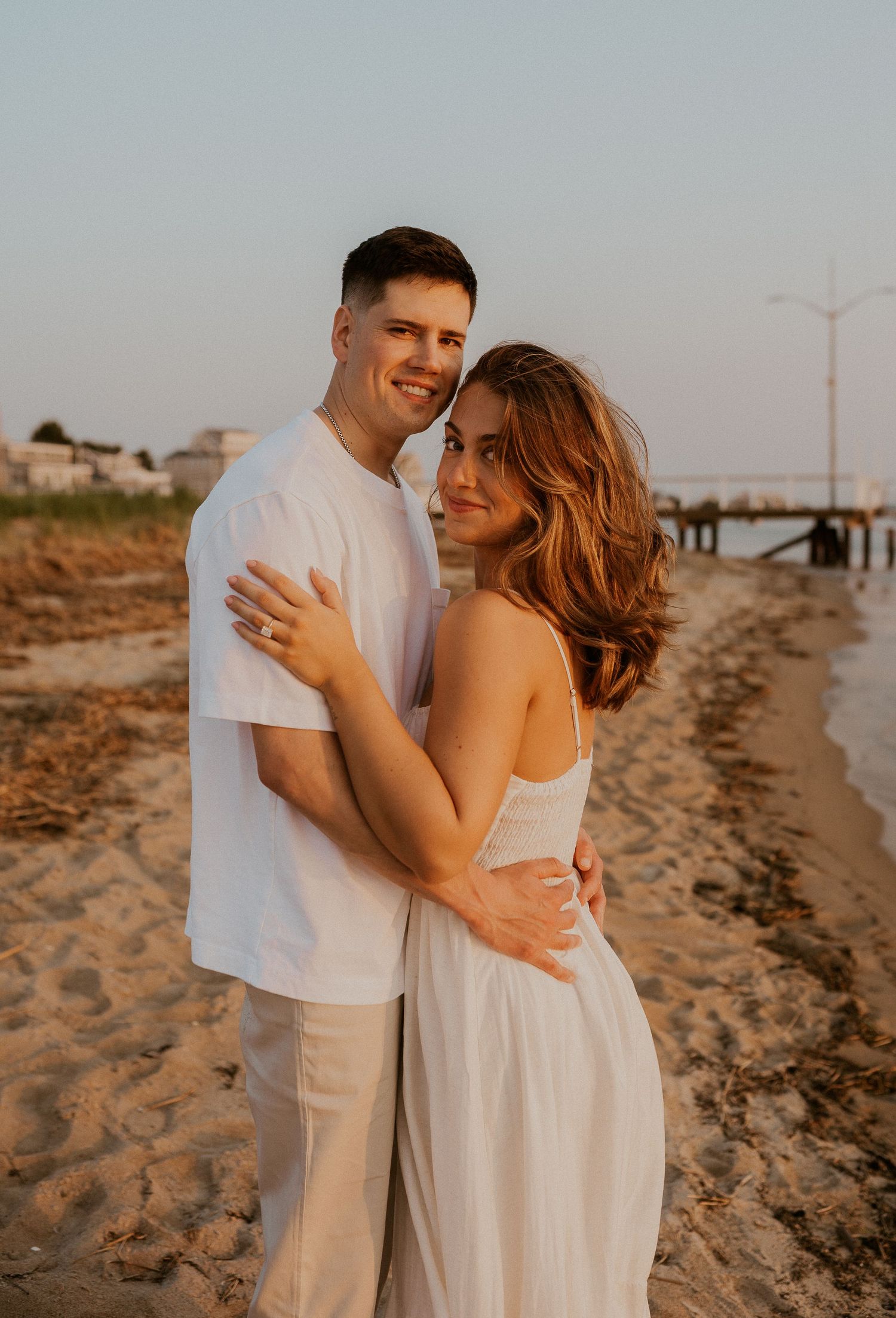 Sunset beach portrait of couple in white outfits sharing joyful moment near pier.