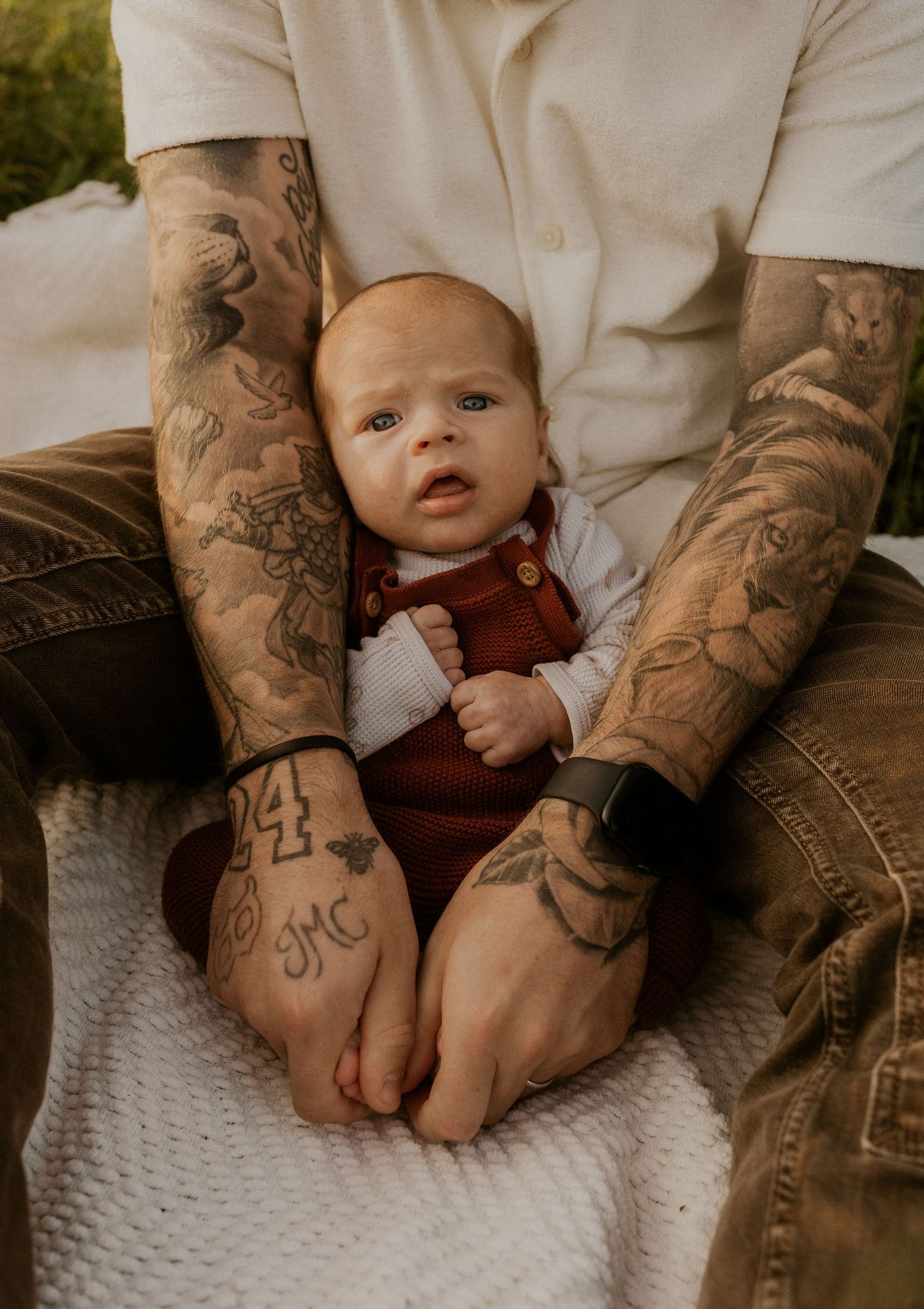 Detailed shot of tattooed hands gently cradling a baby wearing brown clothing.