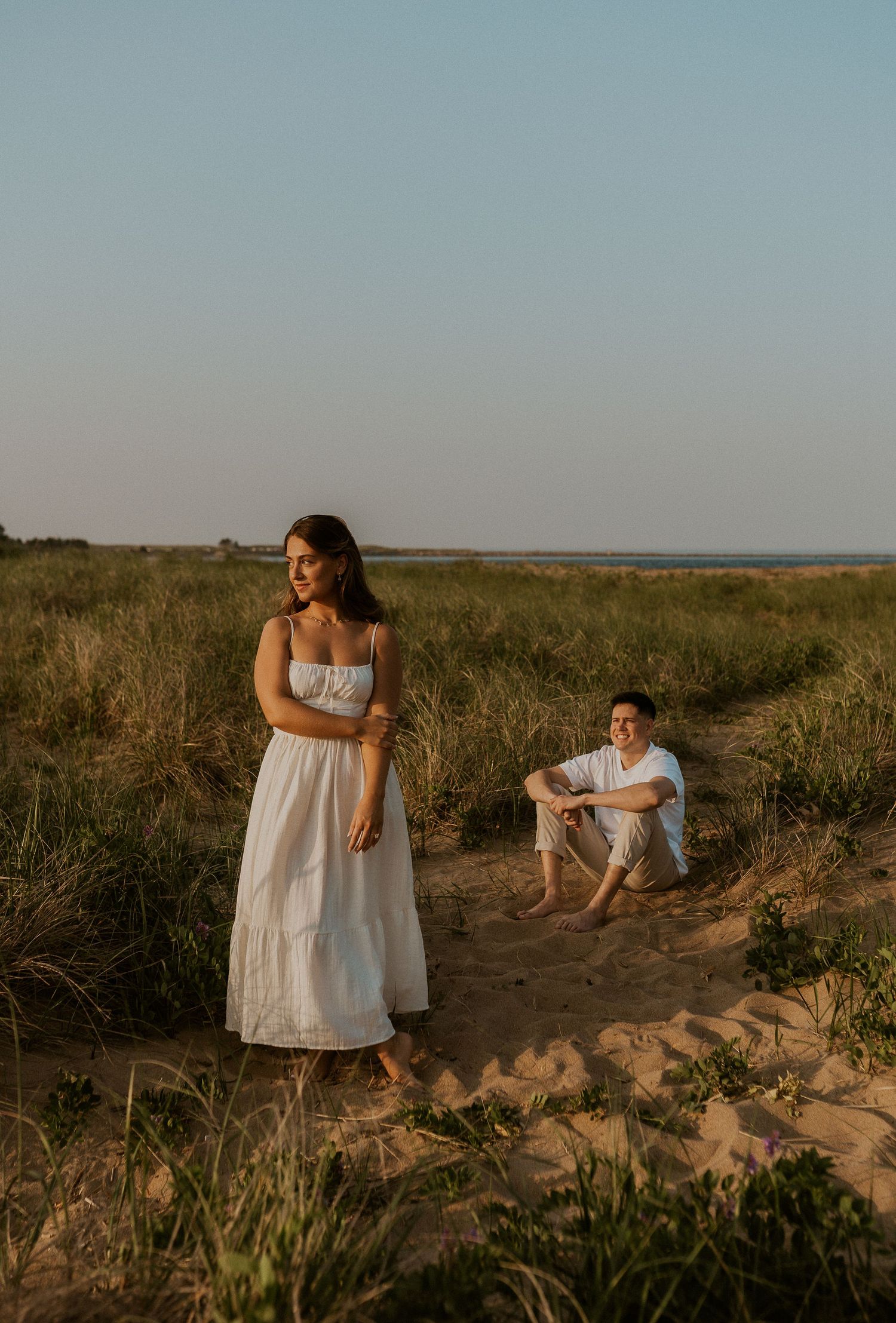 Romantic couple in white outfits posing at sunset on coastal dunes.