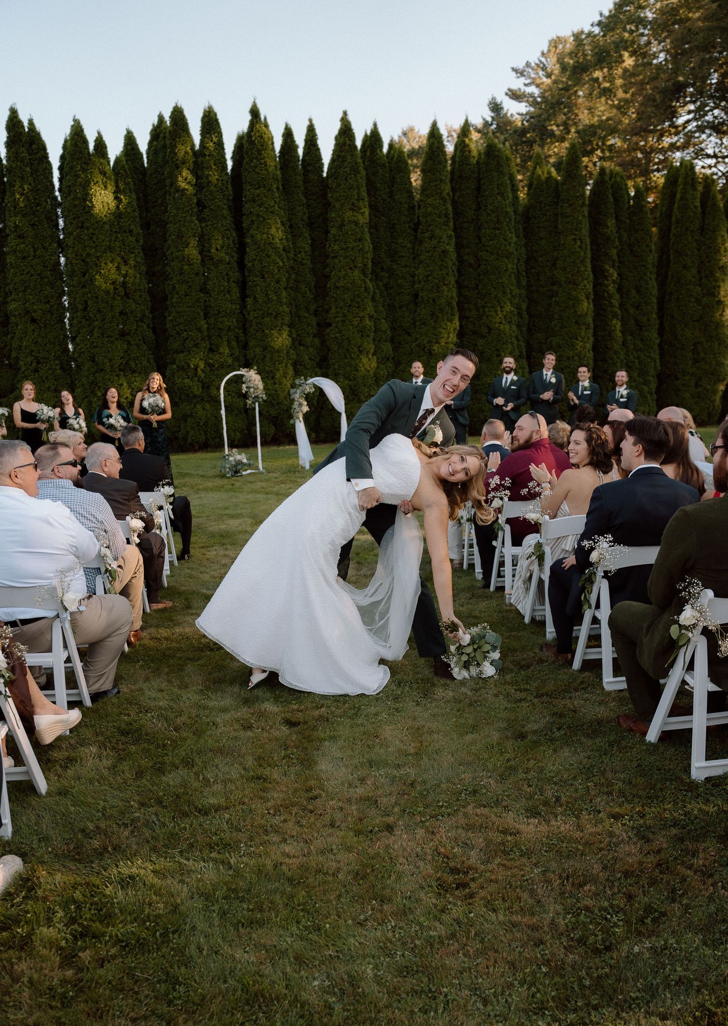 Wedding guests seated in white chairs during an outdoor ceremony with cypress trees forming a natural backdrop.