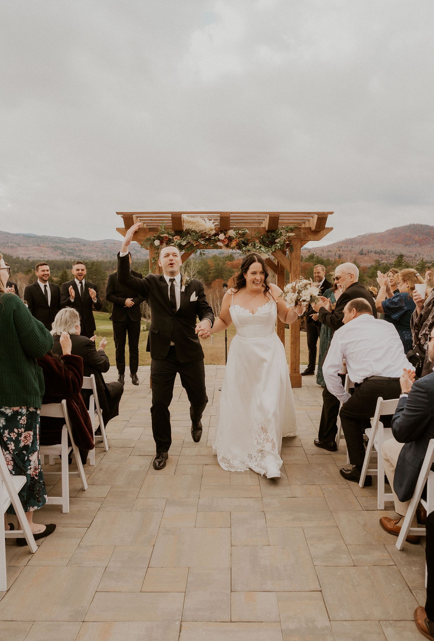 Wedding ceremony guests gathering on a stone patio with wooden pergola and mountain views in the background.