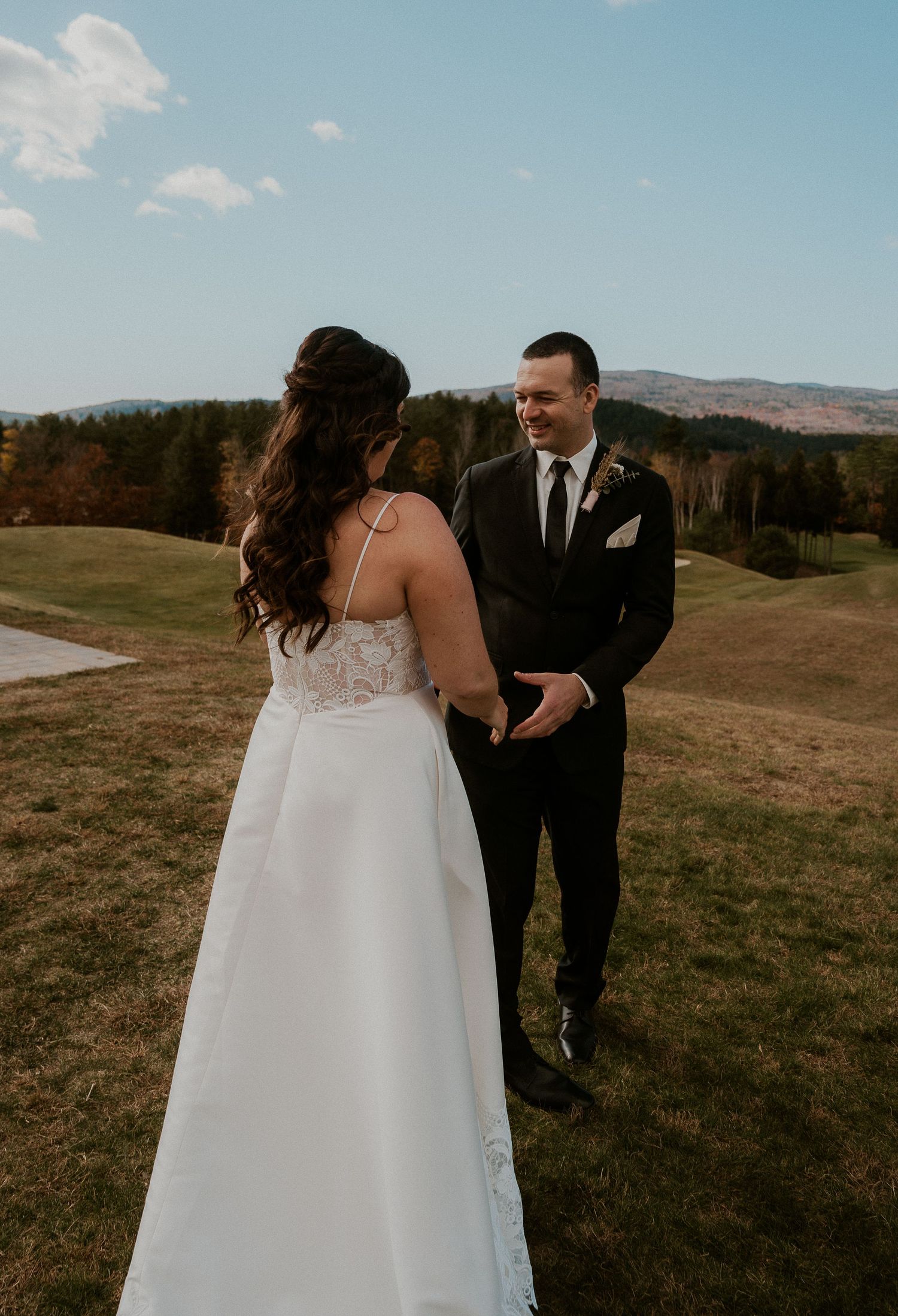 A couple in wedding attire stands together in a scenic mountain field on an autumn afternoon.