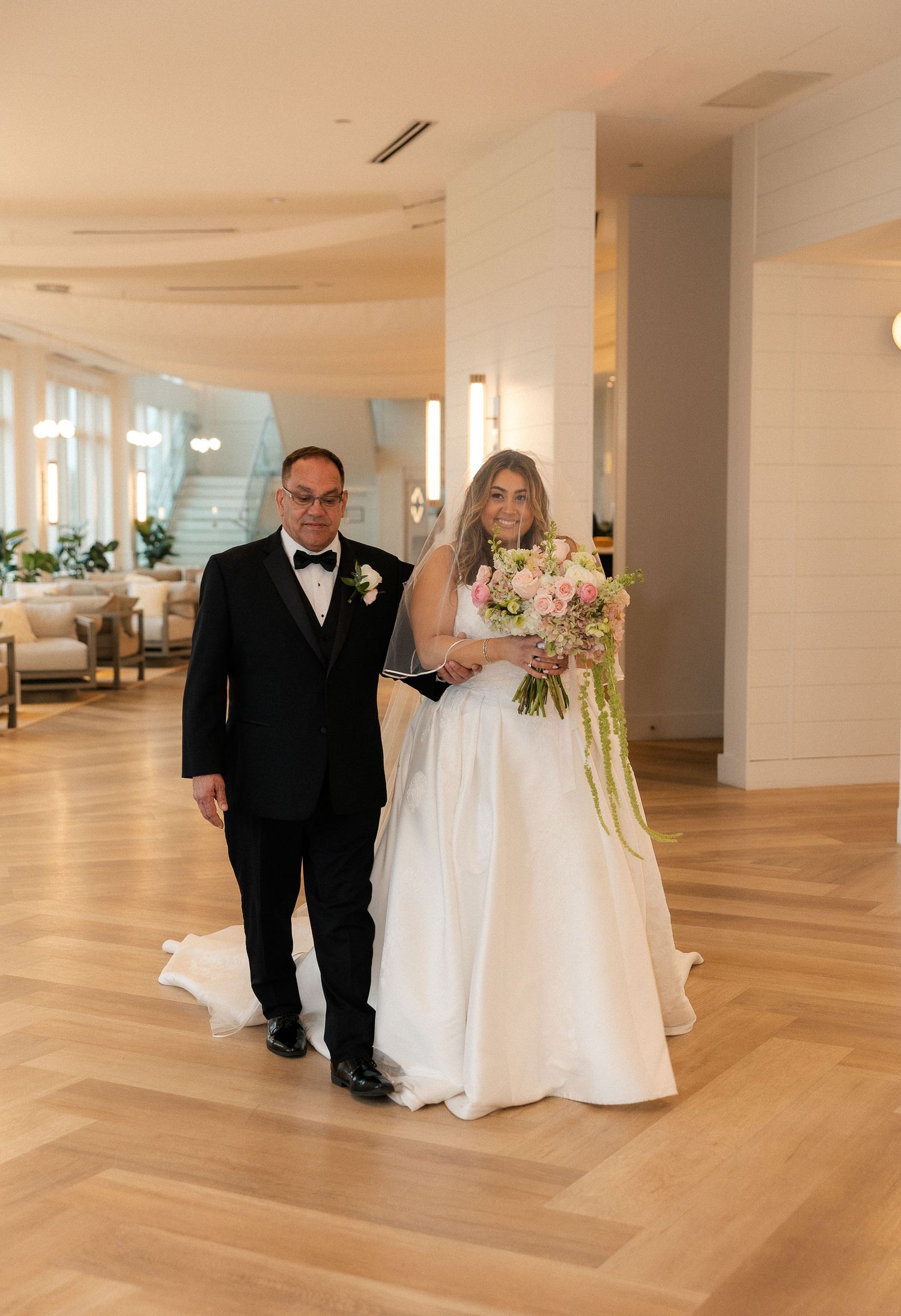 Bride in white ball gown holding pink bouquet walks with escort through elegant hotel lobby on wedding day.