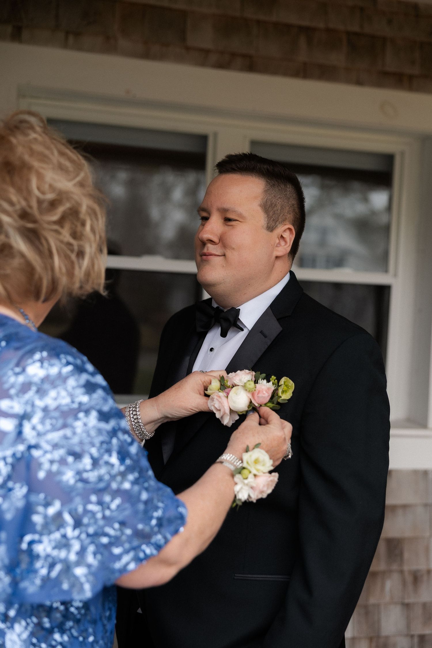 Woman pinning a floral boutonniere on a groom in a black tuxedo outdoors before the wedding ceremony.