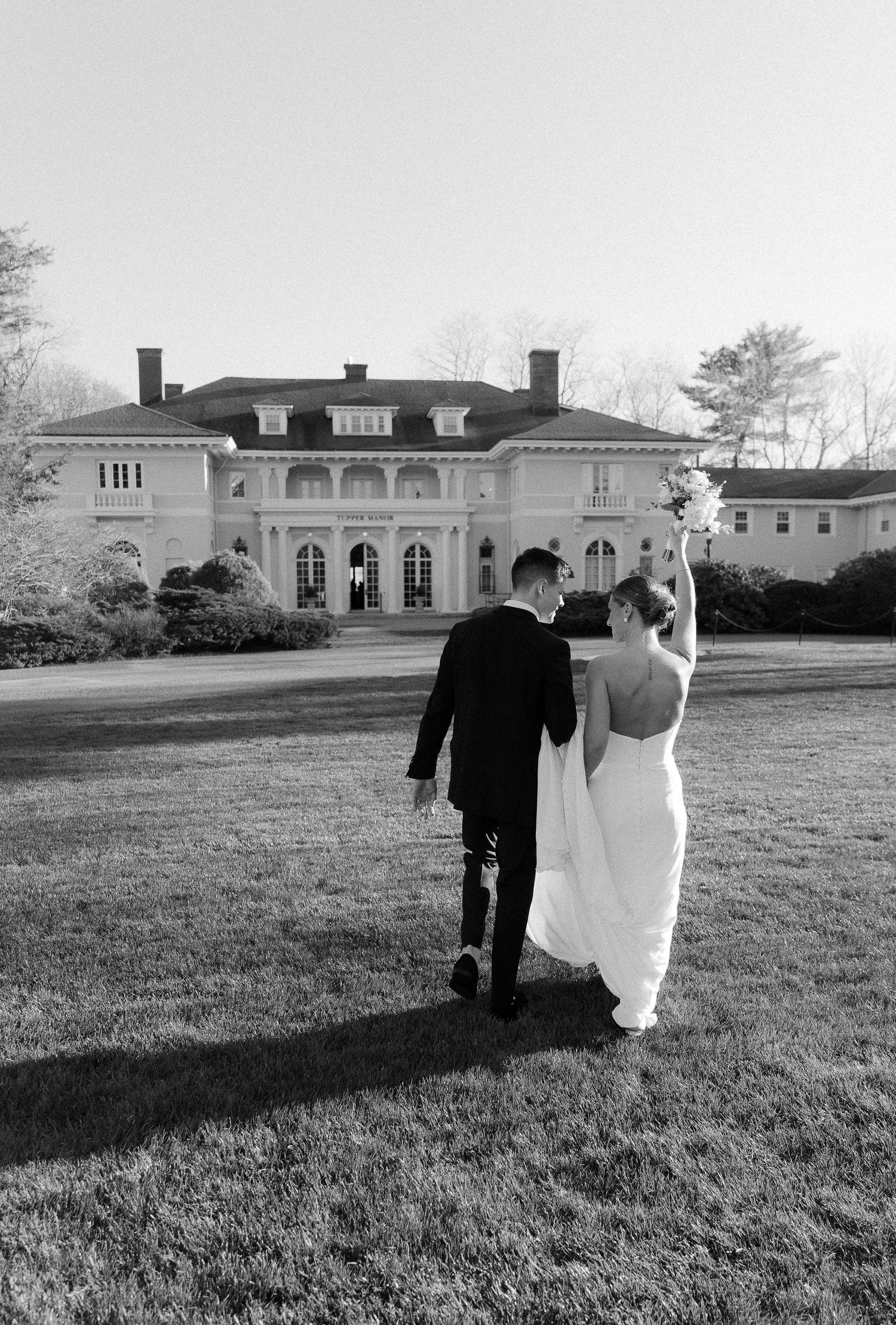 Bride and groom walking toward elegant mansion venue, bride raising bouquet in black and white wedding photo.