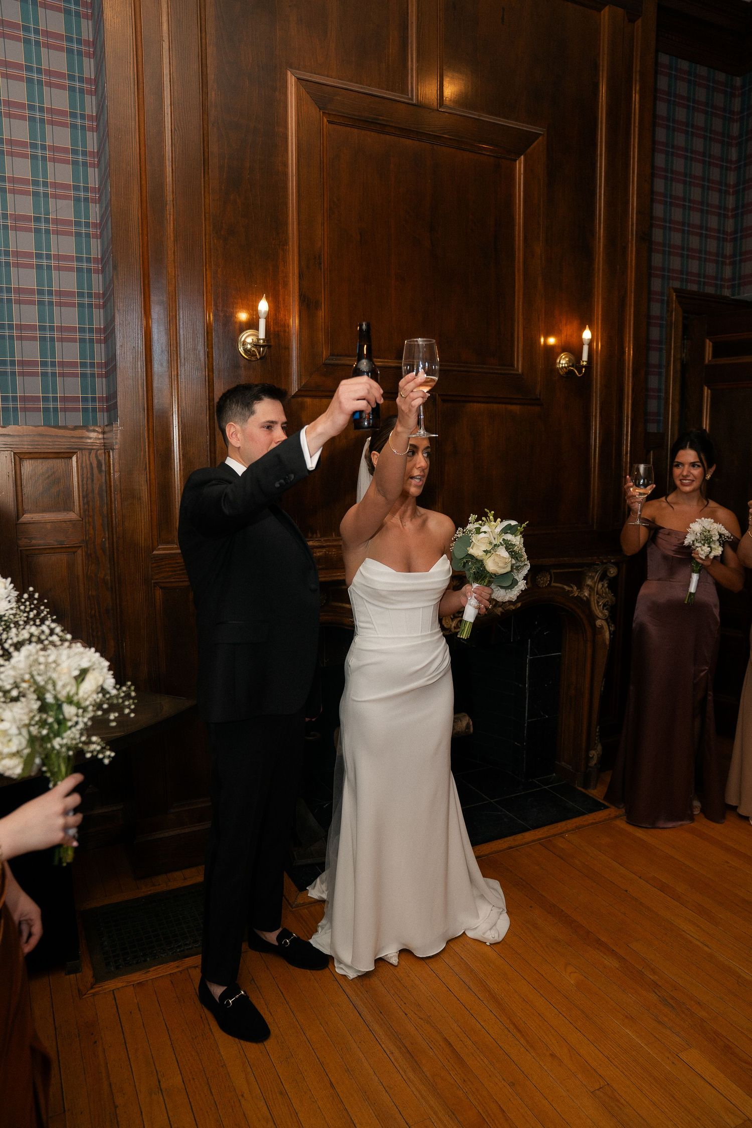 Bride and groom raise champagne glasses in a toast surrounded by wedding party in a wood-paneled reception room.