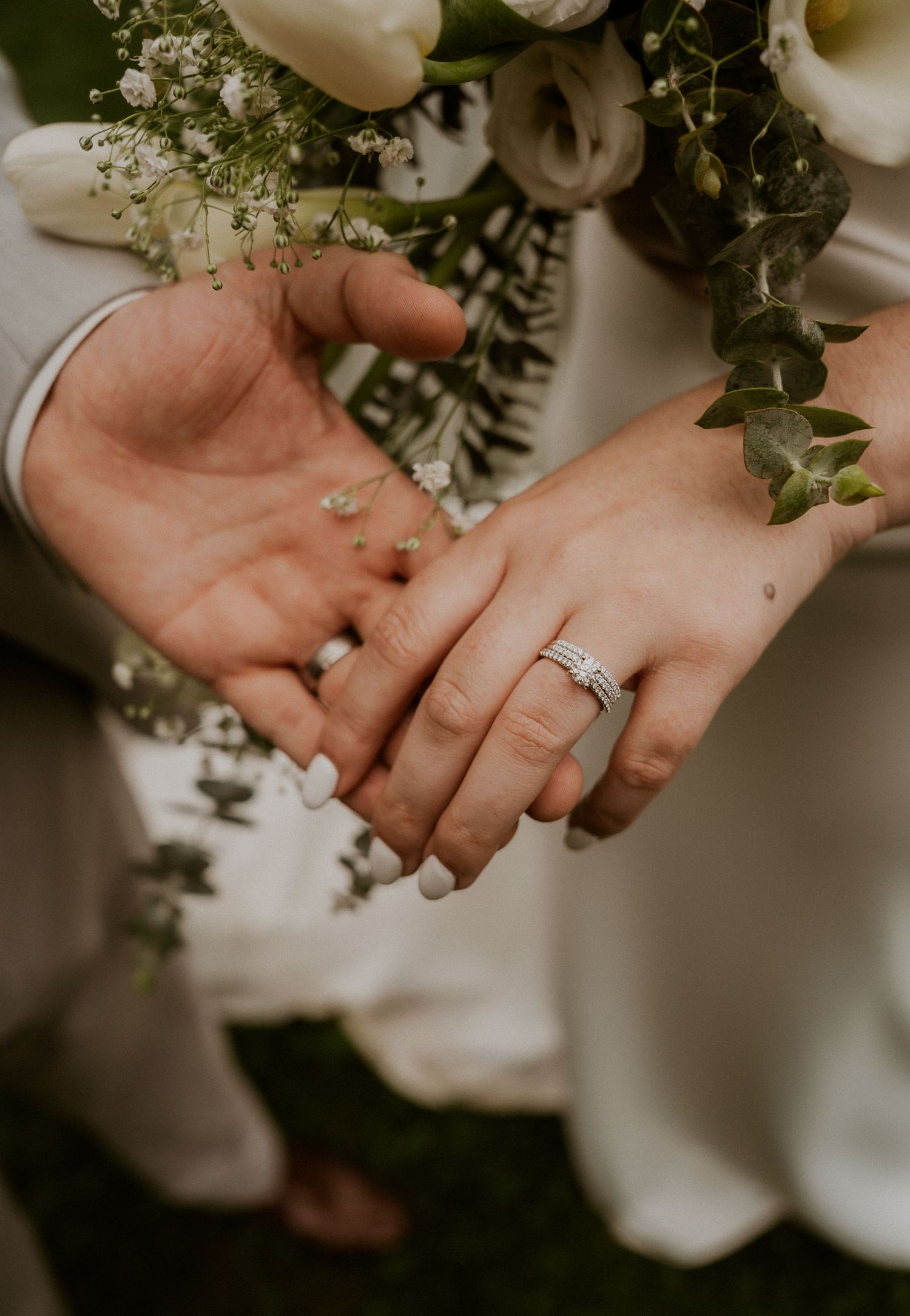 Close-up of newlyweds holding hands, showcasing wedding rings, with a white floral bouquet in the background.